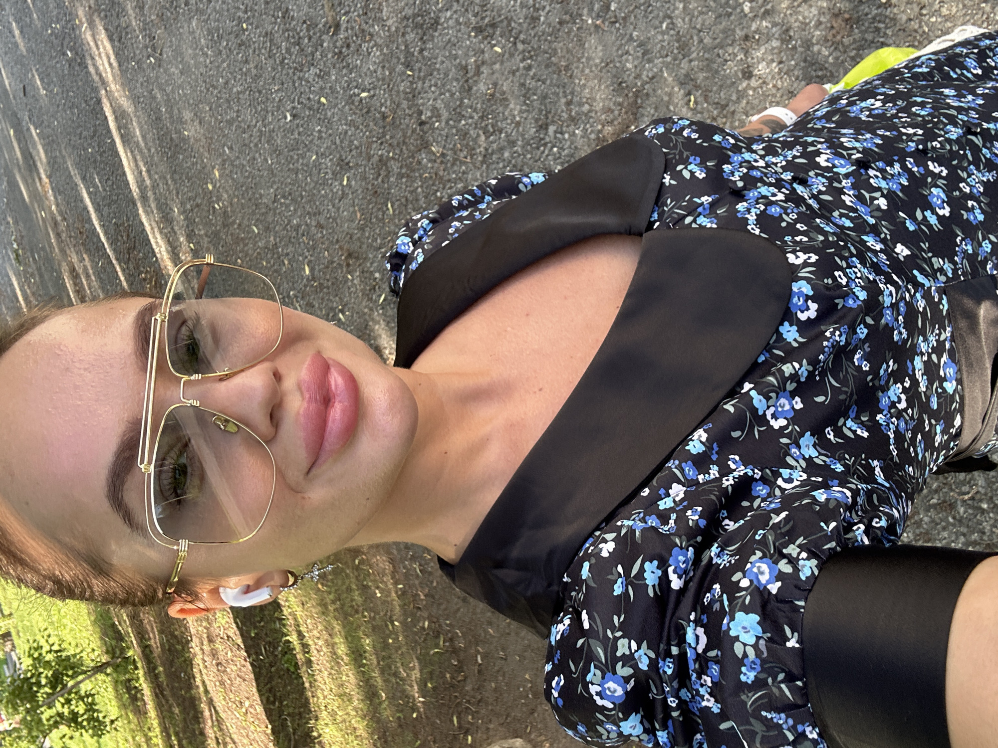 The image shows a young woman with full lips, wearing large gold-rimmed aviator sunglasses and white wireless earbuds. She has a dainty cross earring in her left ear. She is wearing a black dress with a floral pattern featuring blue and white flowers. The dress has a prominent black collar. The background appears to be an outdoor setting with a paved or gravel path and some greenery, possibly a park or a street in the city of Duvankul', Russia. The lighting suggests it is daytime, with dappled sunlight.