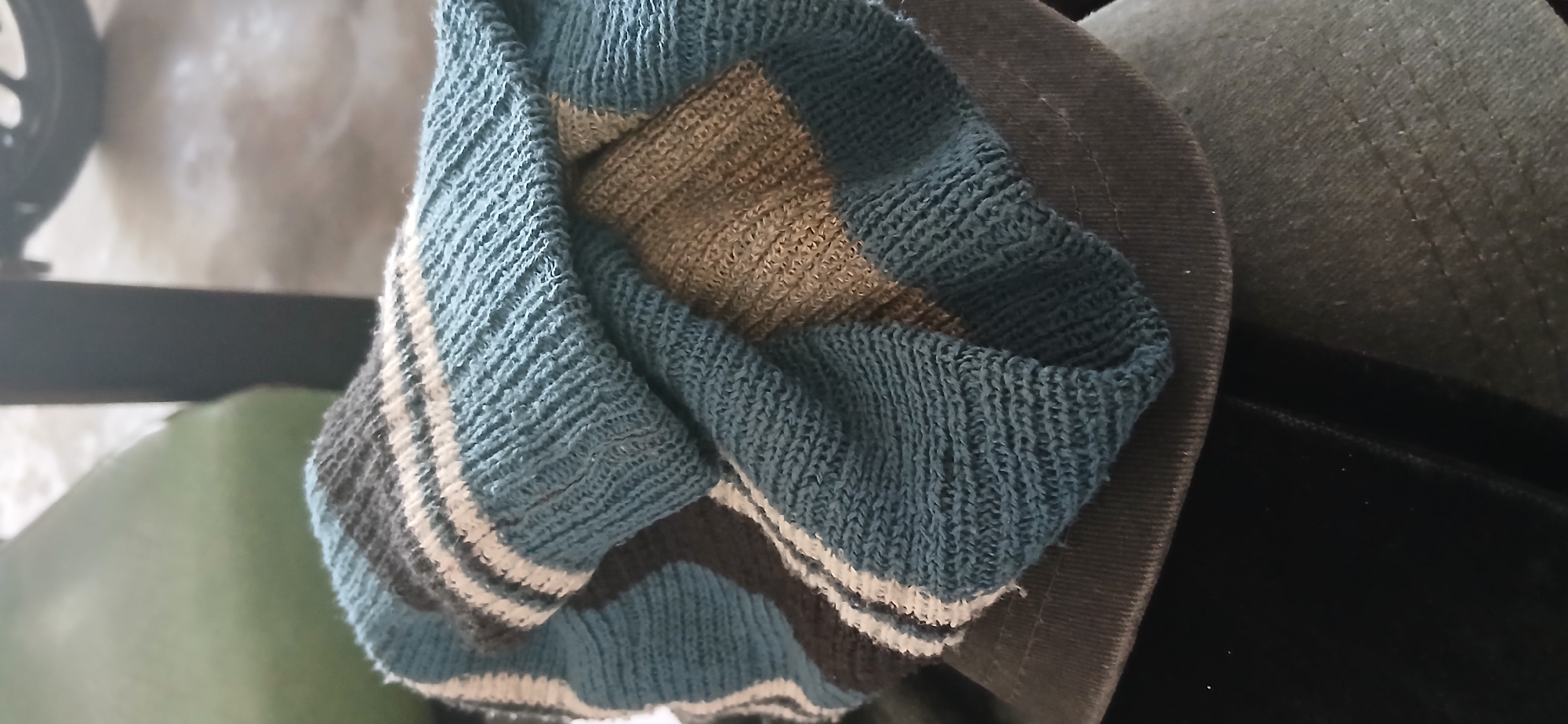 The image shows a close-up of a folded, knitted blue and white striped sock, likely a men's or unisex sock, resting on a dark gray hat with a subtly textured brim. The sock has a ribbed texture and is folded over itself, creating layers and shadows. The hat appears to be made of a fabric like canvas or twill. The background is blurred but suggests an indoor setting with a hint of a greenish object on the left and a dark vertical element on the right, possibly furniture or part of a structure. The lighting is diffused. The image is taken from a slightly elevated perspective, looking down at the objects. There are no people or animals visible. The scene is static and appears to be a casual, everyday setting. The location is identified as Kademangan Kulon, Indonesia.