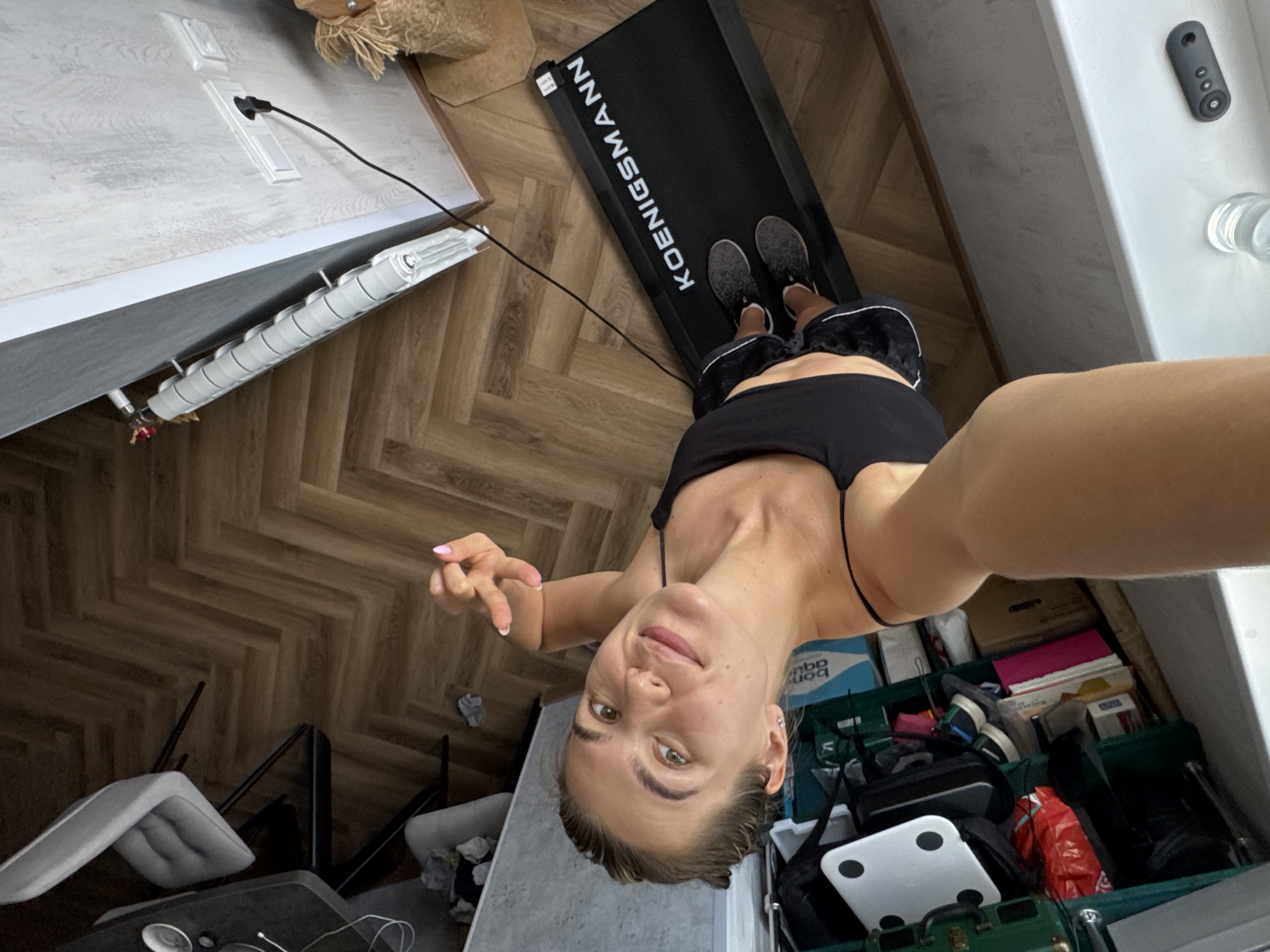 This media file is a selfie taken by a young woman indoors. She is wearing a black sports bra and black shorts. She is standing on a black treadmill with "KOENIGSSMANN" written in white letters on it. Her legs and feet are visible on the treadmill, and she is looking towards the camera. The woman has blonde hair and appears to be in good spirits.
The setting appears to be a room with wooden floors. To the left of the treadmill, there is a white radiator connected to a white electrical outlet. To the right of the treadmill, there is a white surface with a black device and a clear glass with liquid. In the background, there are various objects, including boxes, bags, and a white scale. The lighting suggests it is daytime. The overall scene conveys a sense of a home gym or a personal workout space.