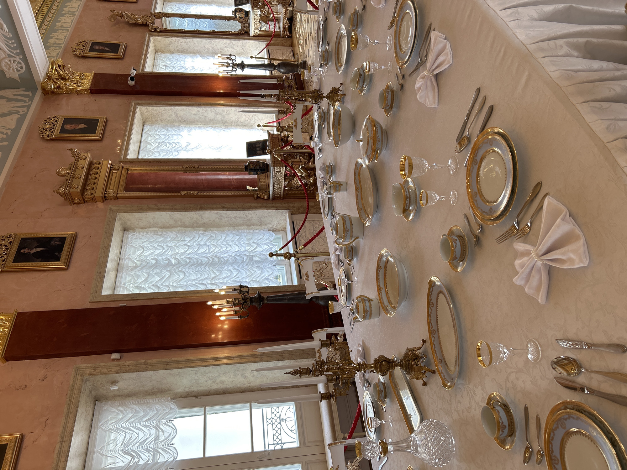 The image depicts a formal dining table set in an opulent room, likely within a historical palace or museum in the city of Dobasnyanski Sel'ski Savet, Belarus. The long table is covered with a white, intricately patterned tablecloth and is meticulously set for a meal. Each place setting includes multiple plates, silverware, a goblet, and a teacup with a saucer. Decorative gold-rimmed porcelain dishes, including bowls, tureens, and serving platters, are arranged across the table. A large, ornate brass candelabra with unlit candles stands prominently near the center of the table, and another candelabra with lit candles is visible in the background. Crystal decanters and glassware are also present. The room itself features high ceilings, large windows draped with white, pleated curtains, and is adorned with classical-inspired architectural elements, including gilded columns and decorative moldings. Framed portraits hang on the walls, and red velvet ropes are used to cordon off areas of the room. The overall impression is one of grandeur and historical significance, suggesting a setting for a formal banquet or exhibition of fine dining. There are no people visible in the image.