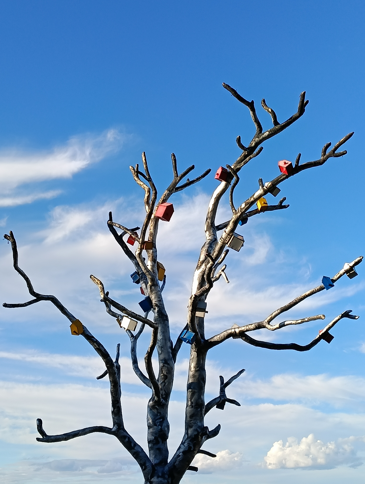 The image depicts a metallic sculpture resembling a leafless tree with multiple branches. Attached to these branches are numerous small, colorful, house-shaped ornaments. The sculpture is positioned against a backdrop of a bright blue sky with scattered white clouds. The base of the sculpture is not visible in the frame. This sculpture is located in Petrozavodsk, Russia, on the Onega embankment. There are no people or animals visible in the image.