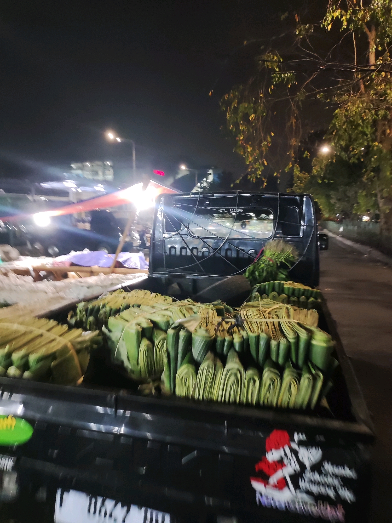 A black pickup truck is parked at night on a street in East Jakarta, Indonesia. The truck bed is filled with bundles of green leaves, likely banana leaves, neatly stacked and tied with twine. In the background, streetlights illuminate a dimly lit market area with tents and other vehicles. Trees with sparse foliage are visible to the right, their branches silhouetted against the dark sky. The overall scene suggests a nocturnal market or delivery operation.