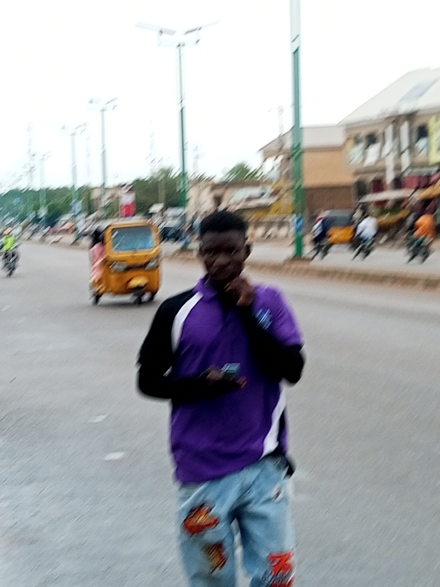 A medium shot shows a young Black male walking towards the viewer on a paved street. He is wearing a purple polo shirt with white and black accents and light blue jeans with red and orange designs. He is holding a dark object, possibly a phone, in his right hand. The background is blurred but shows a street scene with buildings, trees, and several vehicles, including a yellow auto-rickshaw and motorcycles. The scene appears to be in an urban environment, consistent with a street in Gombe, Nigeria, judging by the presence of the auto-rickshaw and the general architecture of the buildings. The sky is overcast.