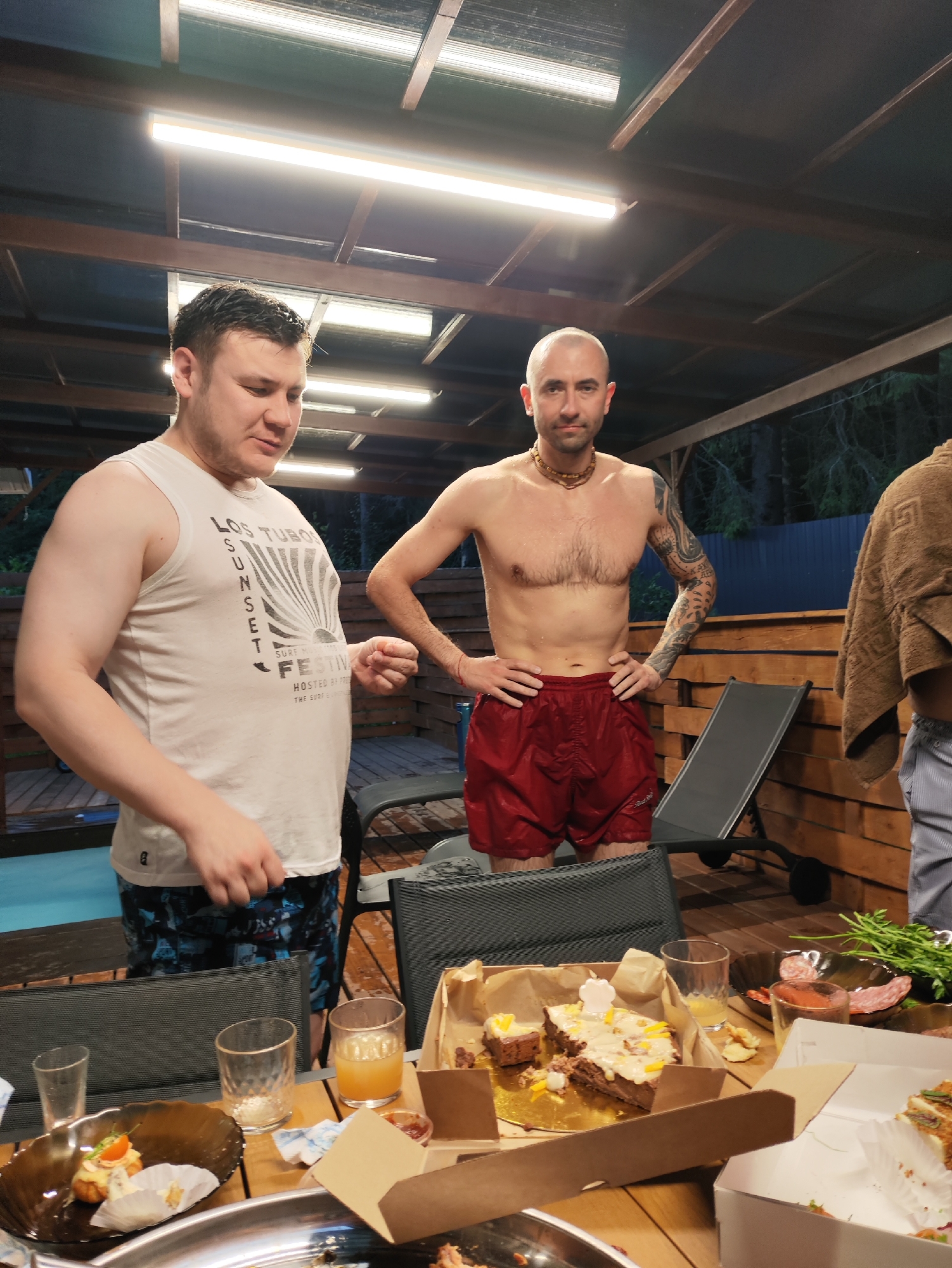 In Старое Мартьяново, Russia, two men are standing on a wooden deck under a covered roof. The man on the left, wearing a white tank top and patterned swim trunks, is looking down and appears to be holding something small in his hand. The man on the right, bare-chested and wearing red swim trunks, has his hands on his hips and is looking at the camera. A table in the foreground is laden with food and drinks, including a cake, slices of salami, and glasses of juice. A towel is draped over a chair in the background, and trees can be seen beyond the deck, suggesting a natural setting. The lighting indicates it is either early morning or late evening.