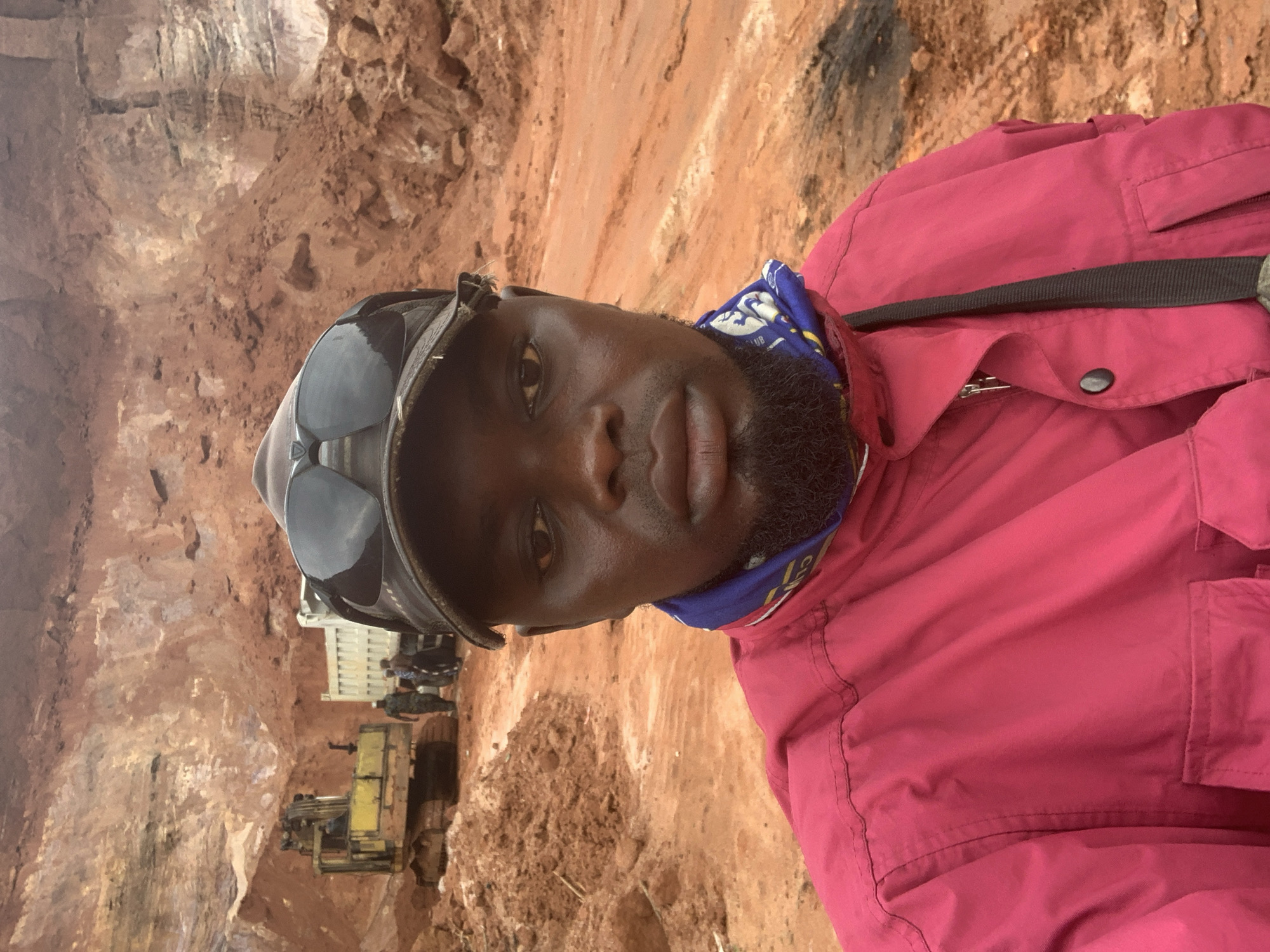 In a quarry in Ajeregun, Nigeria, a selfie is taken by a man wearing a pink jacket, sunglasses perched on a dark cap, and a blue and white bandana. In the background, an excavator is seen next to a large dump truck. Several figures stand near the truck. The environment is characterized by piles of reddish-brown earth, indicative of mining operations.