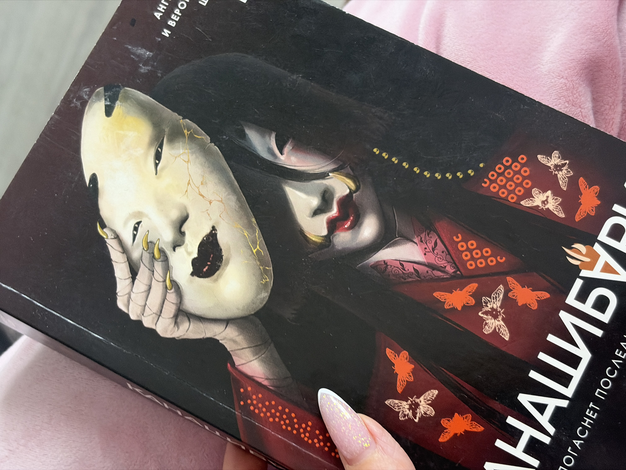 A close-up, oblique shot depicts a book cover featuring a woman with long black hair, red lipstick, and a red kimono, holding a cracked, white mask. The title of the book is "НАШИБУНГ". The photo is taken indoors. There is no indication of Saint Petersburg, Russia.