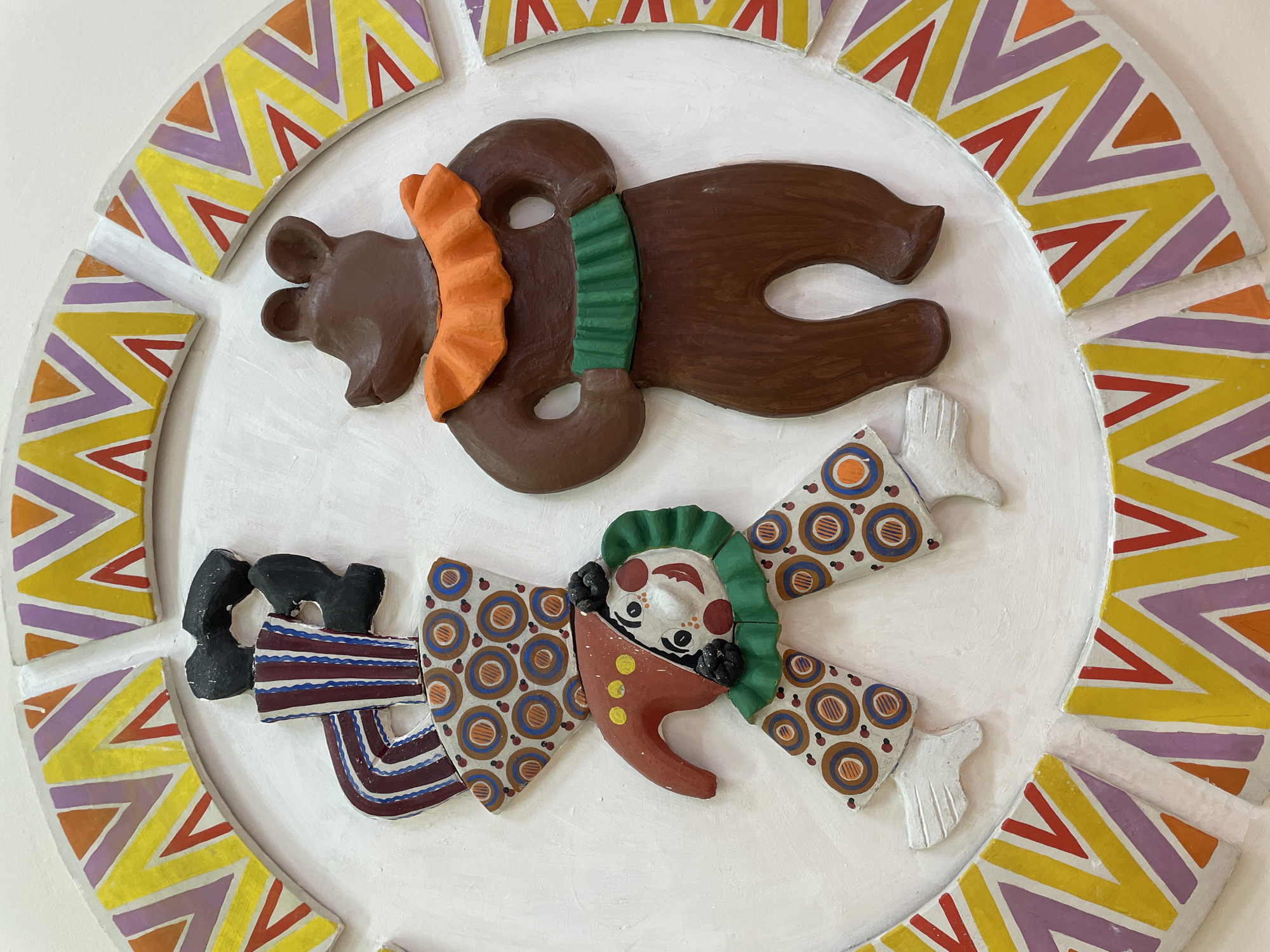 The image shows a relief sculpture affixed to a white wall. The sculpture is circular and appears to be made of painted ceramic or clay. It depicts a bear above and a clown below, both in whimsical, folk-art style.
The bear is brown, with an orange ruffle at its neck and a green ruffle at its midsection. The clown is multicolored with a red, elongated nose, a white face, black hair, and a green ruffle. The clown's clothing is patterned with stripes and circles.
The background of the sculpture is white. Around the edges of the circular sculpture are geometric shapes painted in yellow, purple, and orange.
Based on the context, this could be a decorative element in Izhevsk, Russia.