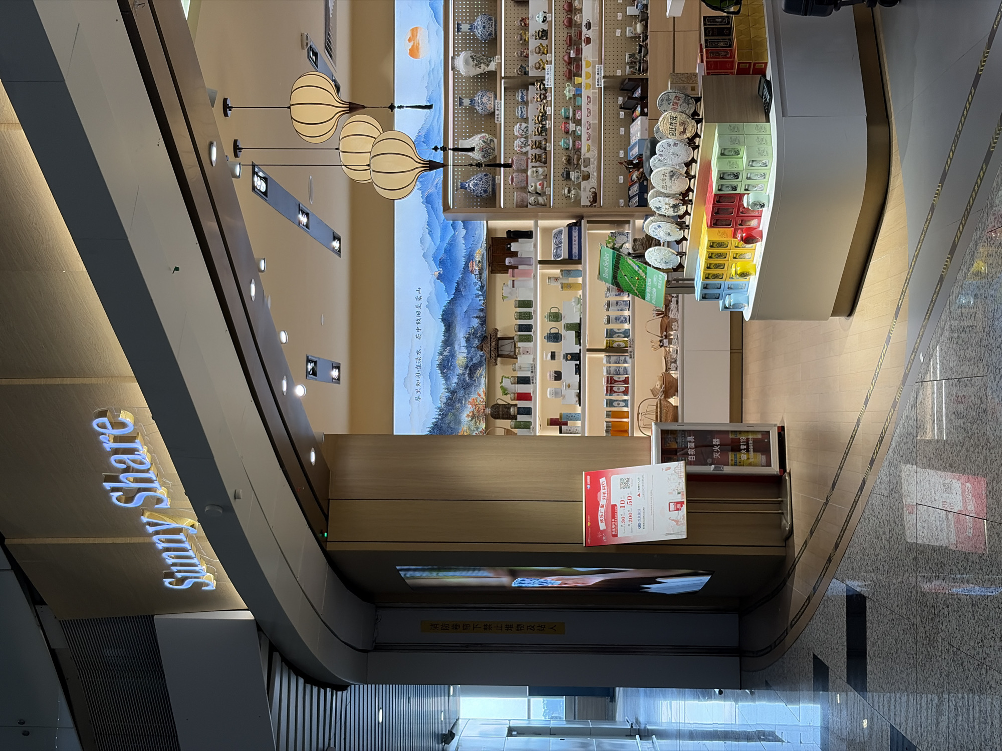 Here is a detailed description of the image:
**Scene:**
The image shows the interior of a store named "Sunny Share," possibly in a shopping mall or airport terminal. The store sells tea and related items, displaying them on shelves against a backdrop of a painted landscape. The perspective is from a slightly elevated position.
**Location:**
The store is located in the Baiyun District, China, based on the context of the image's location.
**Subjects:**
* **Store display:** The shelves are stocked with various teas, tea sets, and other related products.
* **Landscape painting:** A backdrop behind the shelves features a traditional Chinese landscape painting.
* **Signage:** There is signage in Chinese, as well as promotional material related to sales.
**Objects:**
* Shelves filled with tea products.
* Decorative lanterns hanging from the ceiling.
* Signage indicating safety instructions and promotional offers.
* Fire extinguisher and safety mask visible near the counter.
* The "Sunny Share" store name displayed with lights.
**Actions:**
The scene appears static, with no visible human activity. The store is set up for retail purposes.
**Interactions:**
There are no visible interactions in the image.
**Environment:**
The environment appears to be a well-lit, modern interior space with tiled flooring. The design of the ceiling and the curved walls suggests the store is part of a larger, possibly upscale commercial complex.
