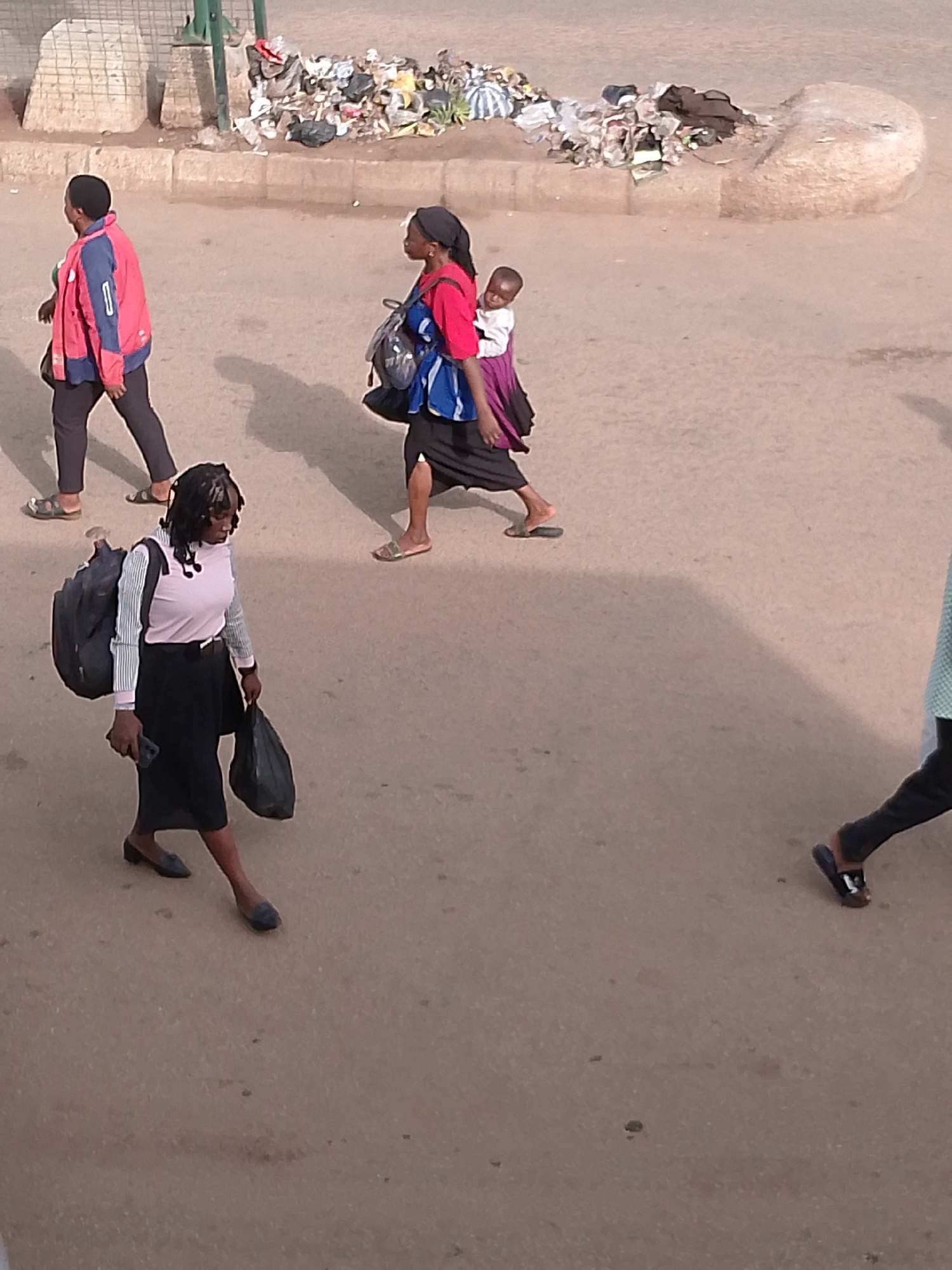 The image depicts a street scene in Bukuru, Nigeria, where several individuals are walking on a paved surface. In the foreground, a woman with braided hair is walking from left to right, carrying a large backpack and a black plastic bag. She is wearing a light pink long-sleeved shirt and a black skirt. To her right, another woman with a headscarf is walking from right to left, also carrying a backpack and a baby strapped to her back. She is wearing a red shirt and a dark skirt. Further to the left, a person in a red and blue jacket is walking away from the camera. In the background, there is a pile of trash on the side of the road, suggesting an urban environment. The ground is a textured, dusty surface, indicative of a dry climate. Shadows are cast on the ground, indicating the presence of sunlight.