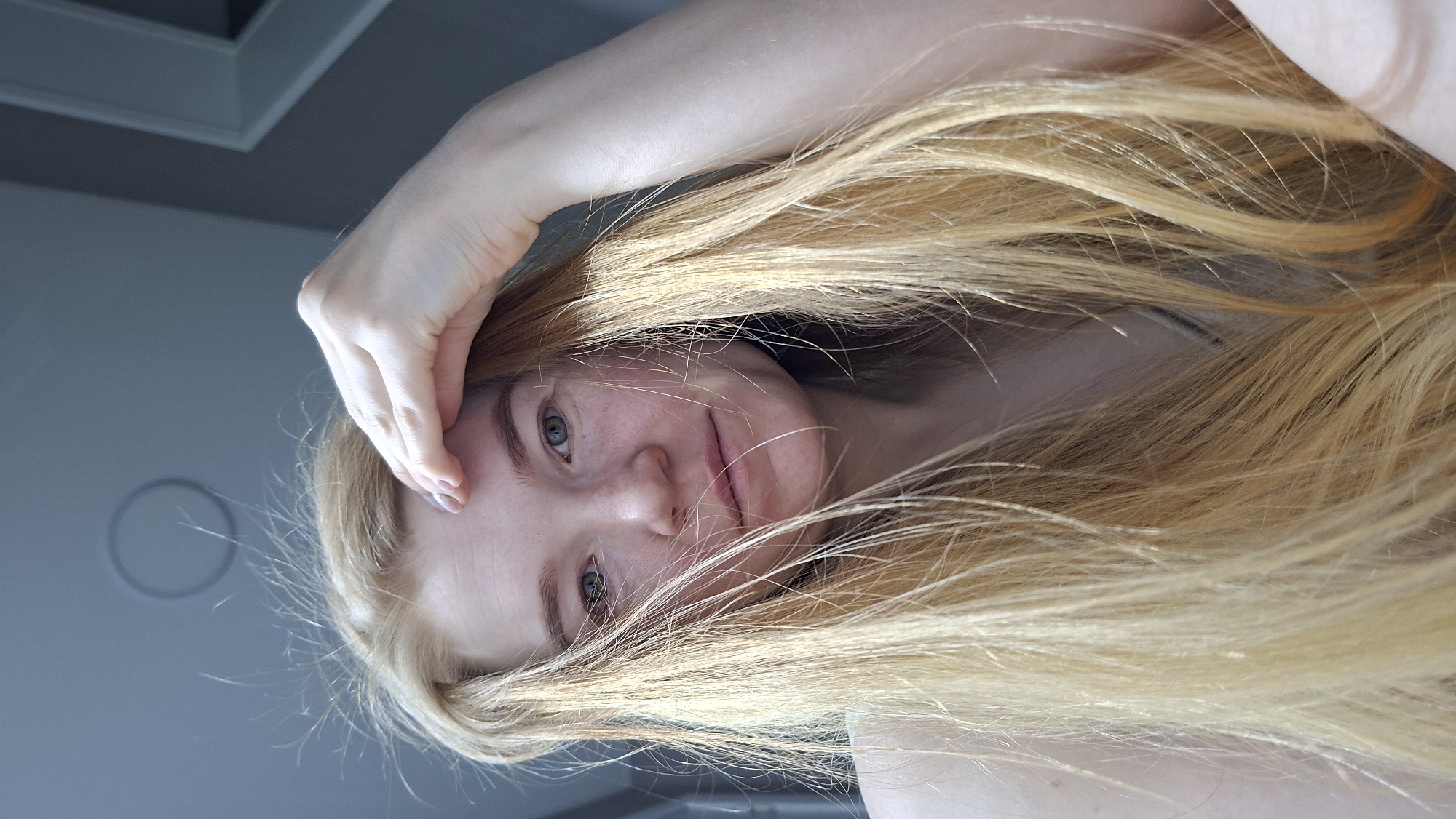 A fair-skinned person with long, blonde hair is positioned in the frame. They are holding their hand near their forehead. Their eyes are visible, and they have freckles on their face. A ceiling and a light fixture are visible in the background. The location in the city of Chaikovsky, Russia is not discernible from the image.