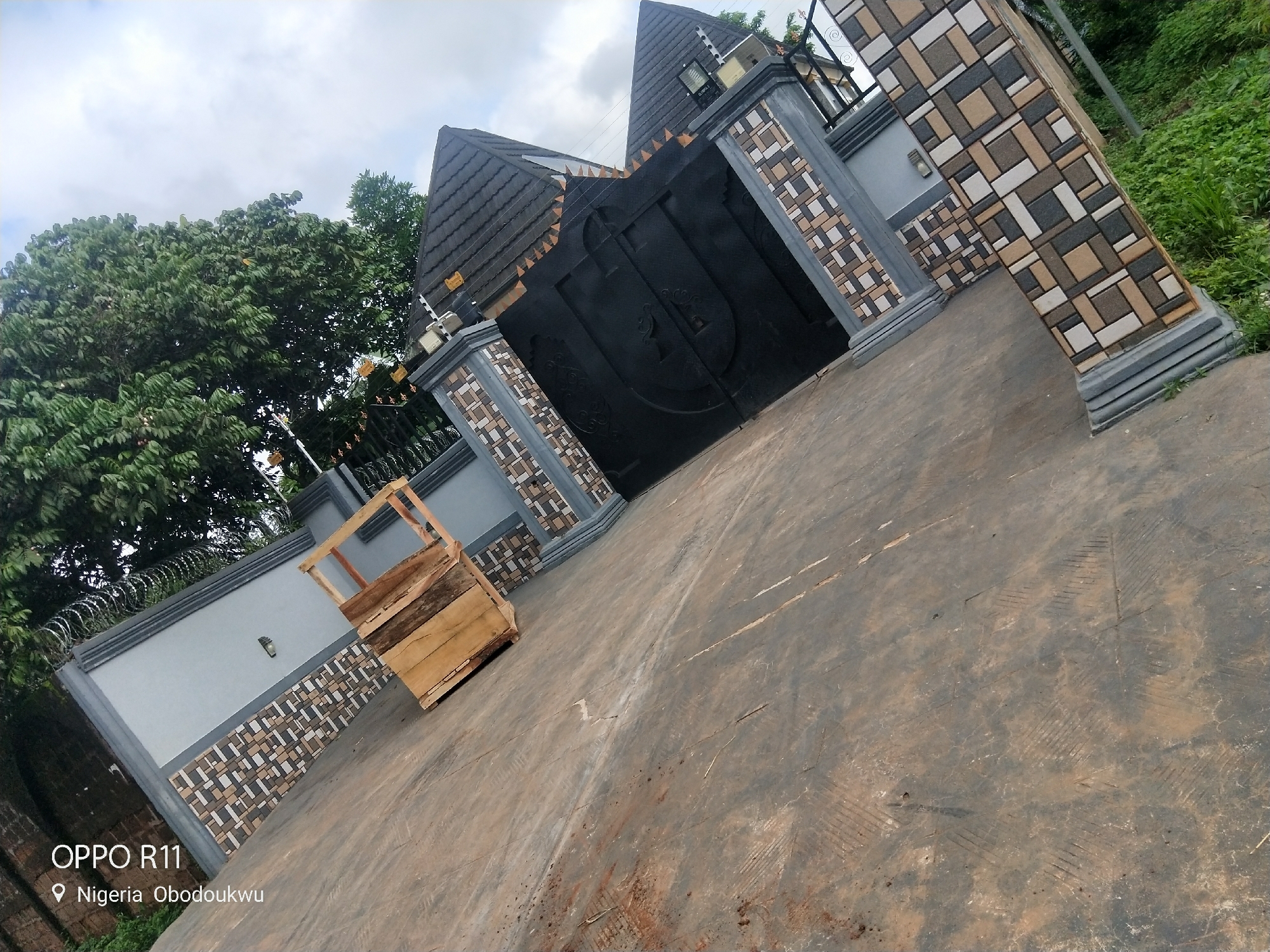 The image shows a large, ornate black metal gate, flanked by decorative pillars. The pillars are made of grey concrete and adorned with a pattern of alternating brown, beige, and grey square tiles. Above the gate, a dark, angled roof with dark grey tiles is visible, with smaller decorative elements and lighting fixtures. To the left of the gate, a grey wall with a similar tiled pattern runs along the property line, topped with razor wire for security. Lush green trees and vegetation are visible behind the wall and to the right of the pillars.
In front of the wall, to the left of the gate, rests a wooden crate. The ground in the foreground is a worn, greyish-brown concrete surface, showing signs of wear and tear with scuff marks and some dirt. The sky above is overcast, with a mix of white and grey clouds, suggesting a cloudy day.
The overall impression is of a secure and well-maintained property entrance in a semi-tropical or rural environment. The location is identified as "Nigeria, Obodoukwu" and the camera information "OPPO R11" is visible in the bottom left corner. No people or specific activities are depicted.