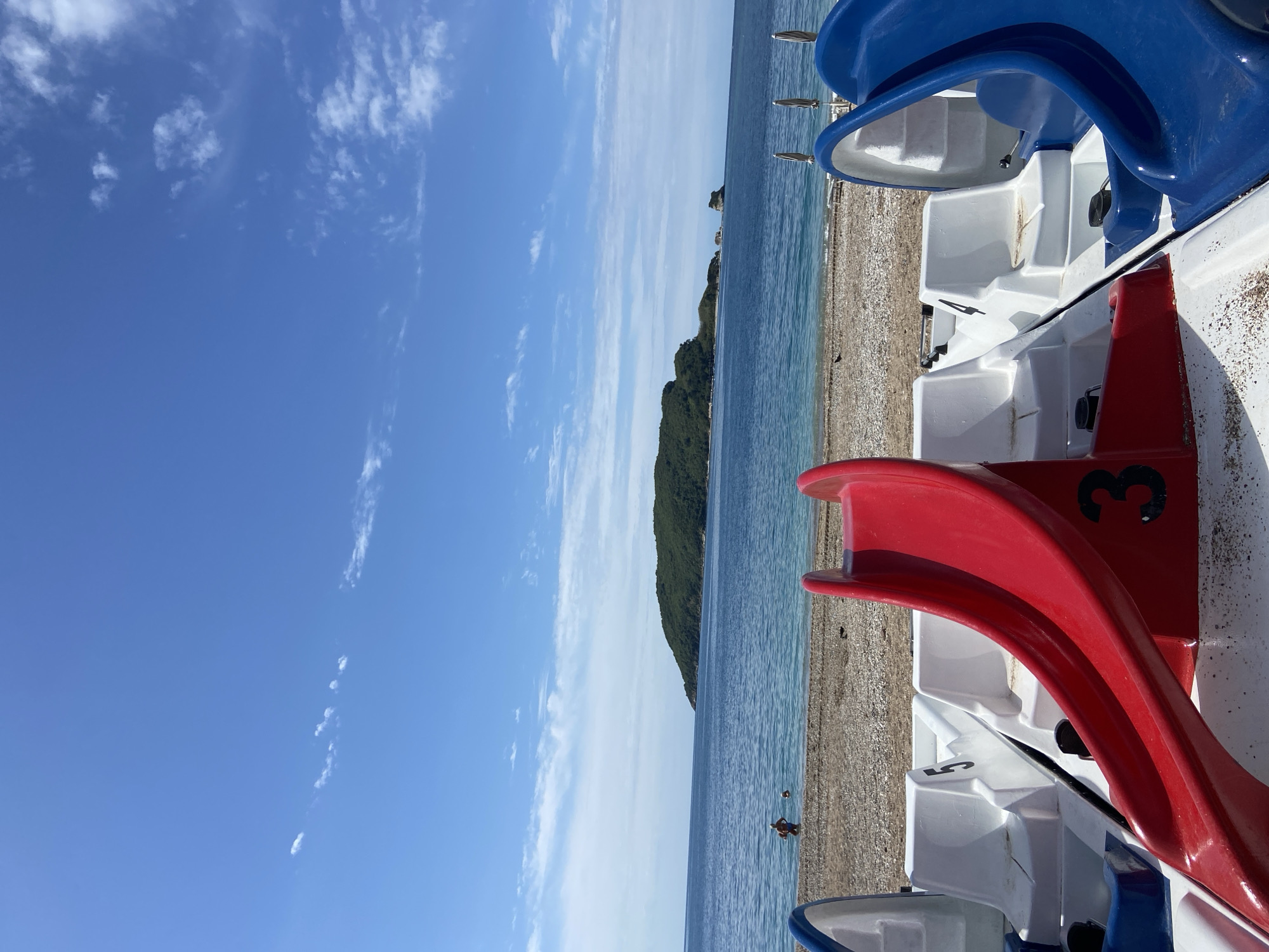 The image shows a beach scene in Budva, Montenegro. In the foreground are several pedal boats, primarily white with red and blue accents. The boats are lined up on the pebble beach. Beyond the beach is the Adriatic Sea, with a green, hilly island visible in the distance. The sky is mostly clear with some wispy clouds. A person is visible wading in the water. The scene appears to be taken on a sunny day.
