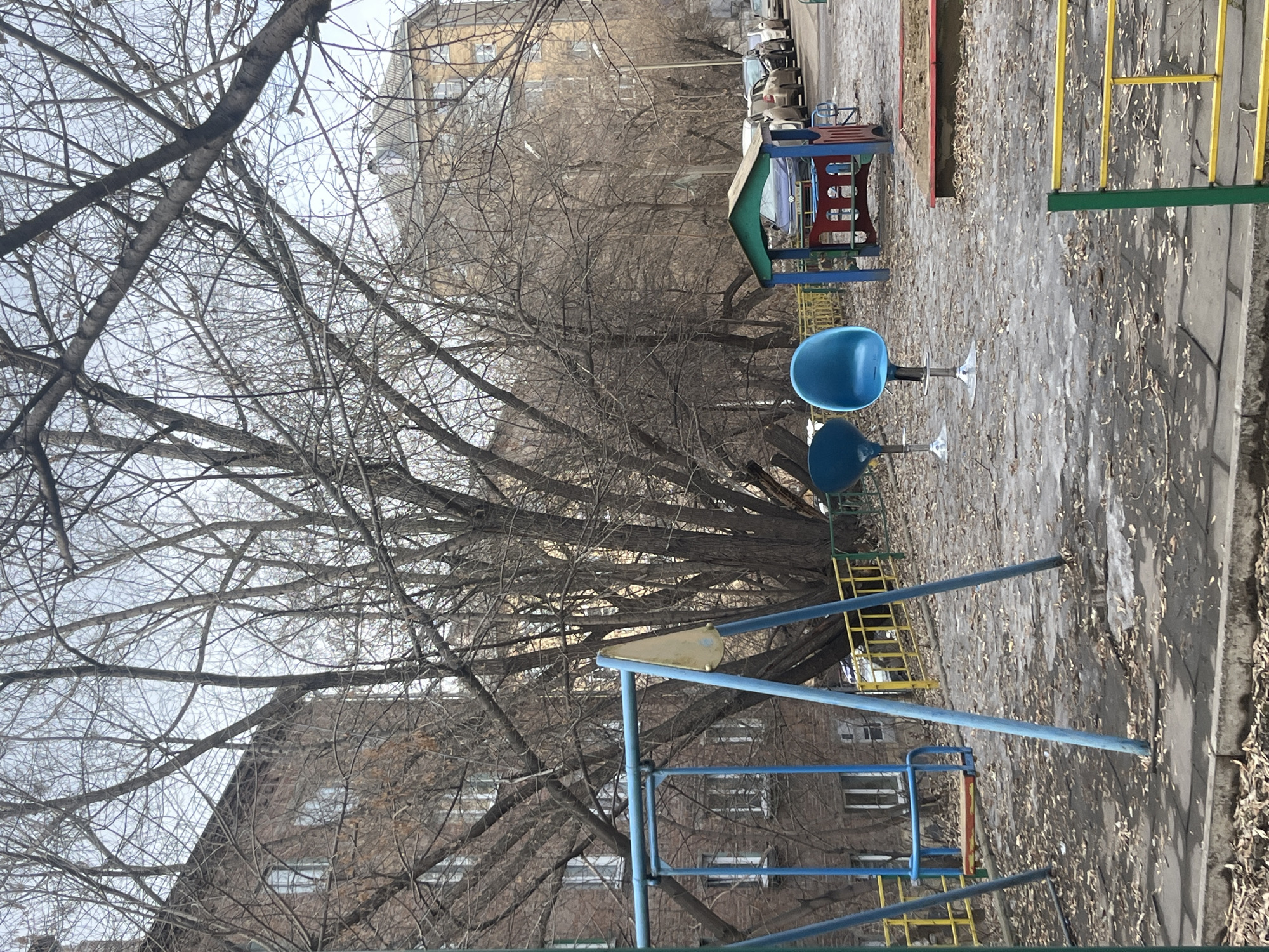 This image depicts an unoccupied residential courtyard or playground, consistent with an urban area in Krasnoyarsk, Russia, during late winter or early spring. The ground consists of dirt mixed with patches of melting ice and scattered dry leaves, bordered by a concrete curb and pathway in the foreground.
In the mid-ground, a blue metal swing set is positioned to the left, featuring two swings: one with a wooden seat painted red and yellow, and another with a yellow triangular seat. Two blue, modern-style plastic chairs with chrome pedestal bases stand centrally on the ground, one notably taller than the other. To the right, a small, multi-colored wooden playhouse with a green roof, blue walls, and a red base is visible, situated next to a sandbox with a red-painted frame. Yellow and green metal railings or fences demarcate various sections of the playground.
The background features numerous deciduous trees with bare branches, partially obscuring multi-story brick residential buildings. On the far right, a line of parked cars is visible on a street or parking lot behind the trees and playground. The sky is overcast. No individuals are present in the scene.