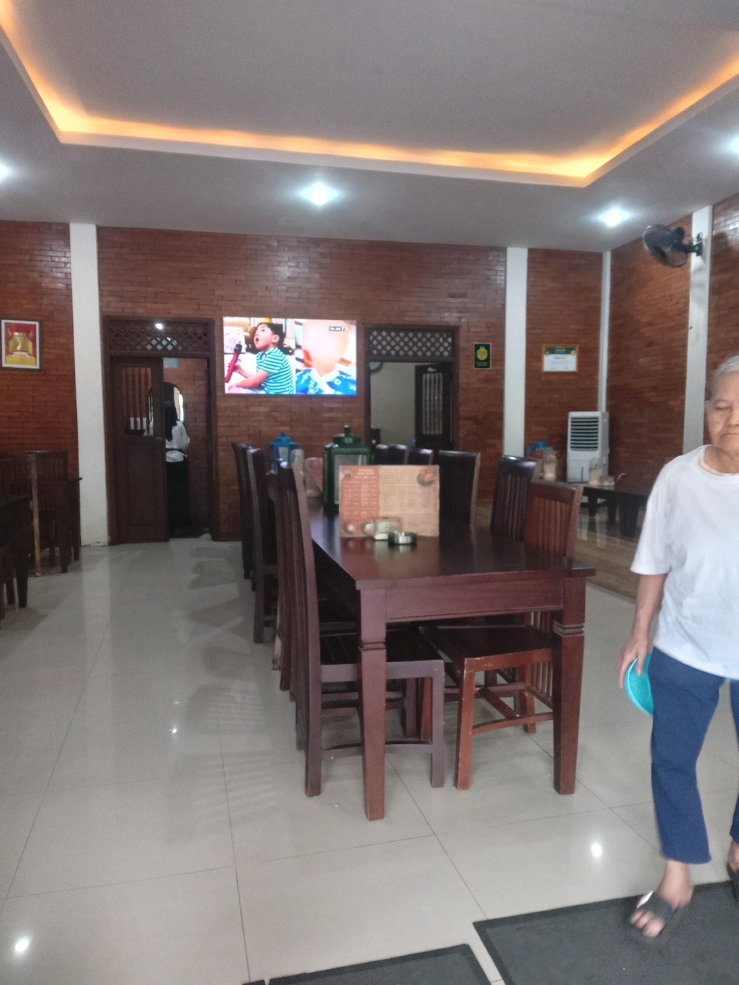 The image shows the interior of a restaurant in Yogyakarta, Indonesia. A long wooden dining table with chairs is centrally located, set against a backdrop of exposed brick walls. On the wall above the table, a flat-screen television displays a video of a child singing. A person in a white t-shirt and blue pants is standing to the right of the table, holding a blue plate. The floor is tiled and appears to be well-maintained. The restaurant has modern lighting fixtures on the ceiling, with recessed LED strips adding ambiance. The overall atmosphere suggests a casual dining establishment.