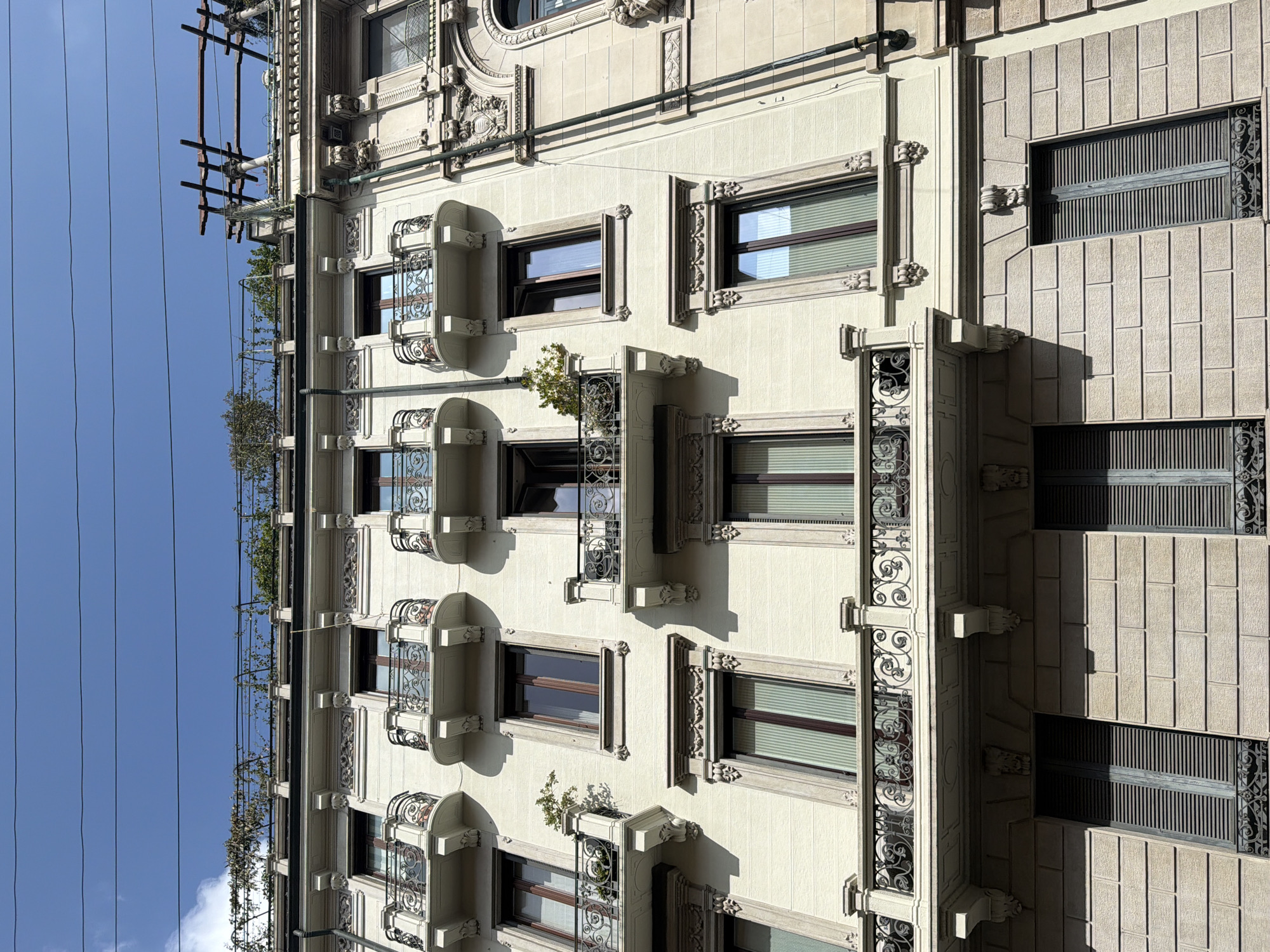 The image depicts the façade of a building in Milan, Italy, notable for its ornate architectural details. The building is characterized by a light-colored, possibly cream or beige, exterior with multiple windows of varying sizes and designs. Some windows are adorned with small balconies featuring intricate wrought iron railings. A vertical row of decorative pilasters runs along one side of the building, adding to its elaborate design. On the upper floors, vegetation grows along the structure, suggesting the presence of a rooftop garden or greenery. A series of overhead wires stretch across the sky, visible on the left side of the frame, indicating an urban setting. The lighting suggests it is daytime, with shadows cast on the building's surface.