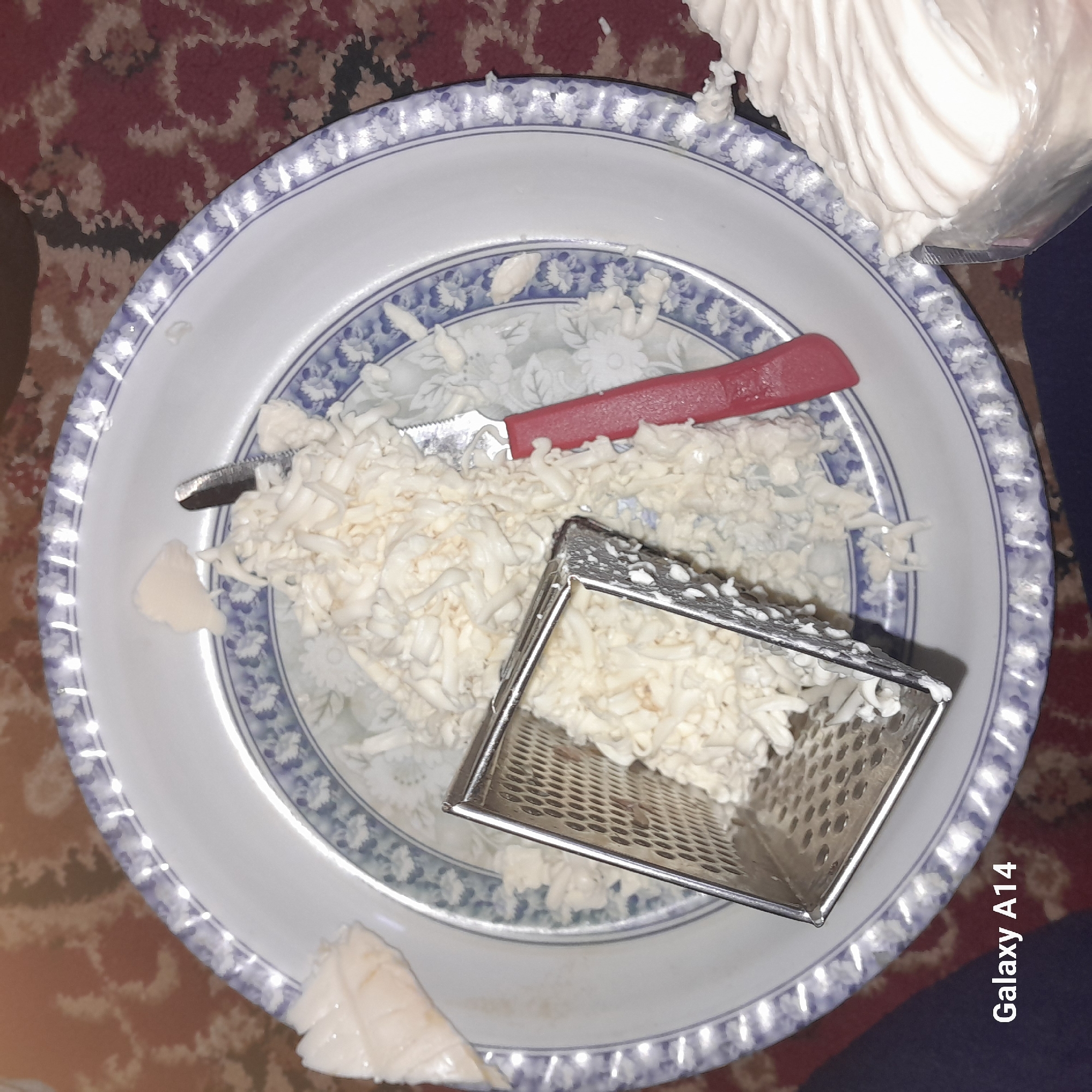 A top-down shot shows a blue and white patterned plate containing shredded white substance, possibly soap or cheese, with a metal grater and a red-handled knife amidst the shreds. A partially visible stack of the same white substance in a plastic bag is in the upper right corner. The background includes a patterned surface, possibly a carpet or tablecloth. The location is Unknown, Unknown.
