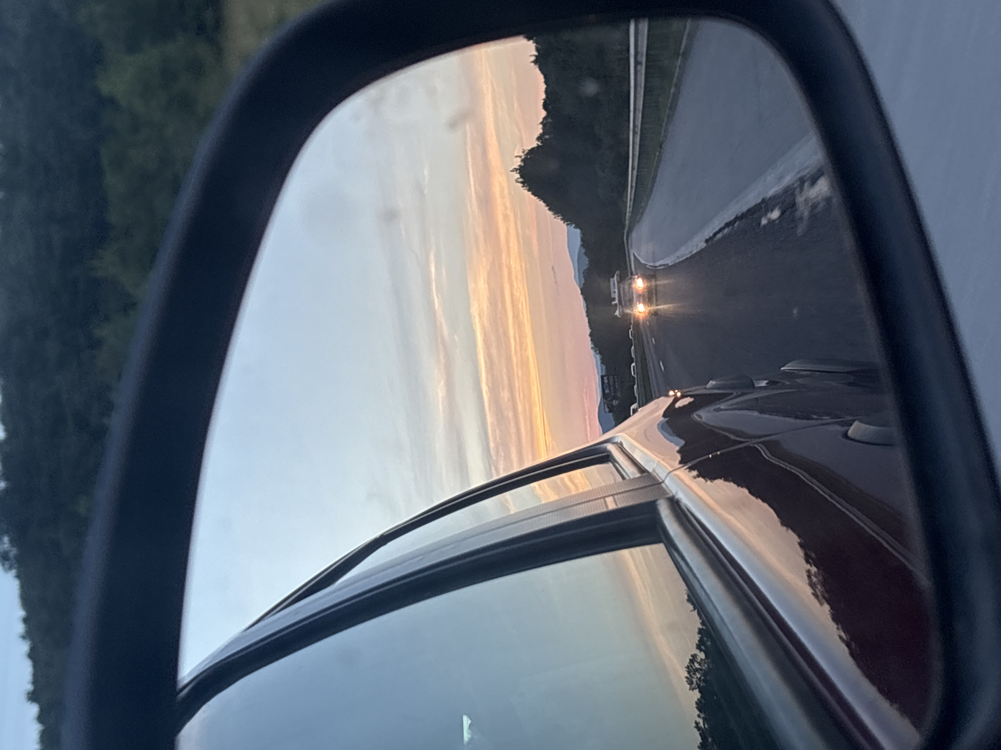 The image shows a view from a car's side mirror, capturing a sunset scene on a highway. In the foreground, the reflection of the car's dark red body is visible, along with parts of its window and frame. The mirror reflects the road ahead, where another vehicle with illuminated headlights is driving. The sky exhibits a warm gradient of colors, transitioning from orange and pink hues near the horizon to lighter blues and grays higher up, indicative of dusk. Trees and hills form the background on either side of the highway. The overall environment appears to be a rural or semi-rural setting, possibly on the outskirts of a town or city. The specific location in Kostadinkino, Bulgaria is not visually discernible from the image itself.