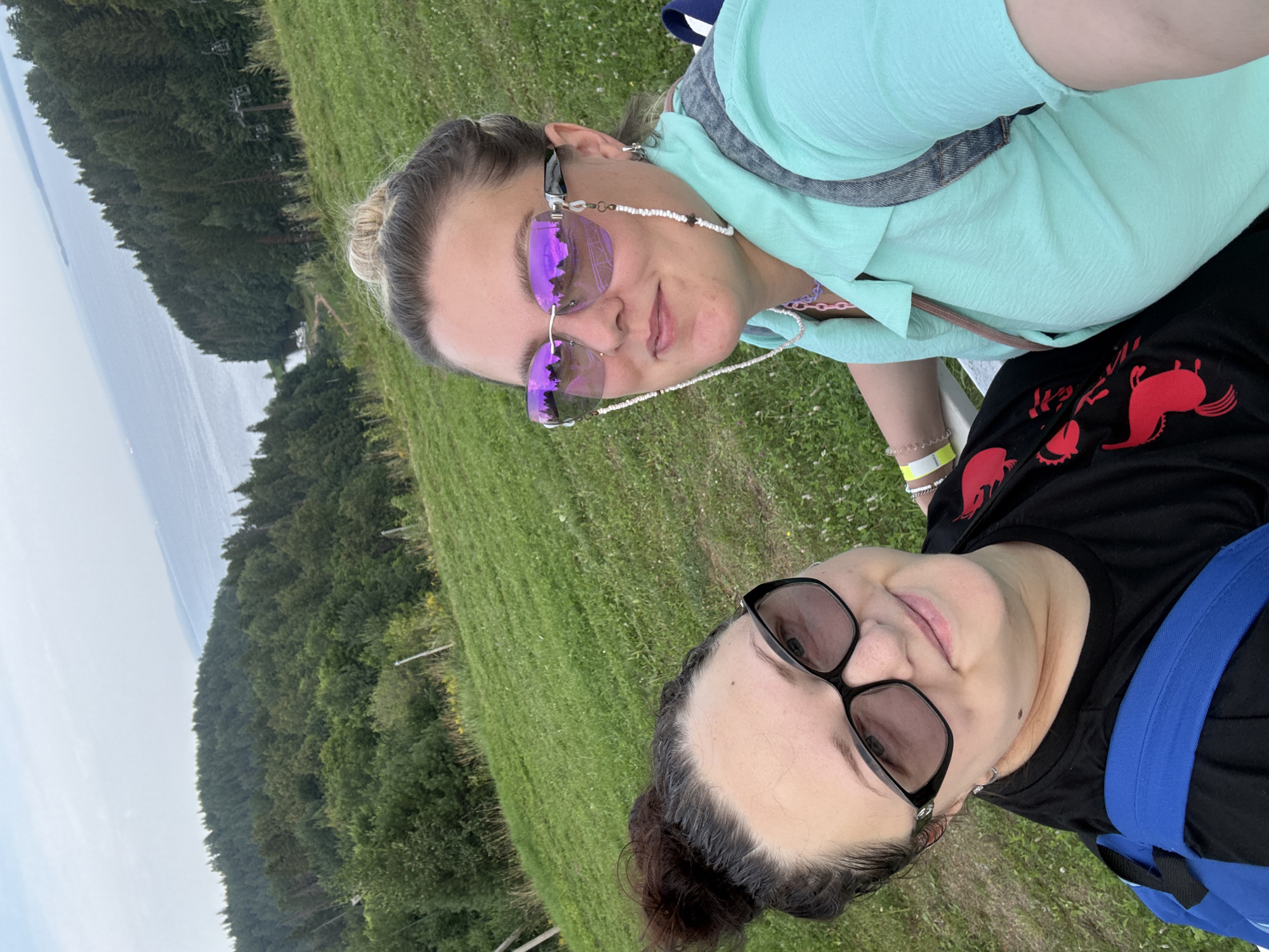 In Galevo, Russia, two women are captured in a selfie on a grassy hill overlooking a body of water and a densely forested area. The woman on the right wears a teal shirt, denim vest, and purple-tinted sunglasses. The woman on the left is in a black shirt with a red graphic and dark-rimmed glasses. Both have their hair up in buns. The environment is a hilly, green landscape with trees and a cable car system visible in the distance. The sky is overcast.