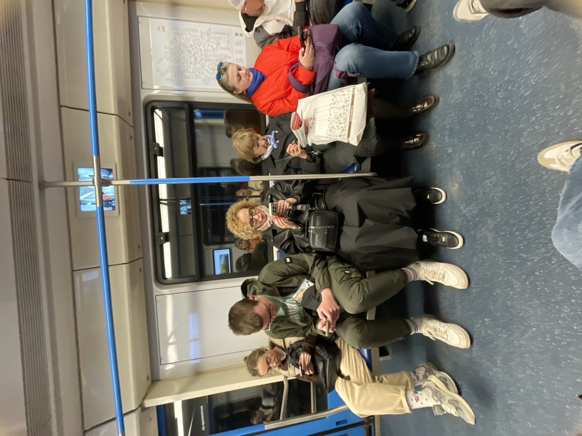 The image depicts the interior of a Moscow metro train carriage. Several passengers are seated along the side of the carriage. One woman wears a bright red jacket. Another is wearing a black coat with curly hair. Two younger individuals are seated near the camera, looking at their mobile phones. The train's interior features a map displayed on the wall. A blue metal support pole runs vertically along the side of the carriage. The floor is blue.