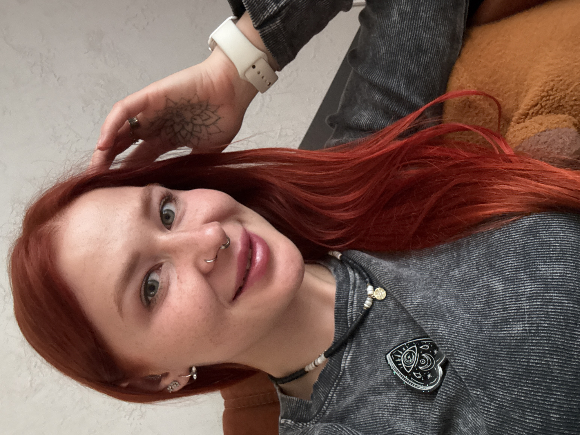 A woman with bright red hair smiles at the camera. She has fair skin, freckles, and green eyes. She has a ring in her nose and earrings in her ears. Her left hand is raised, touching her hair. She has a tattoo on the back of her hand and a white watch on her wrist. She is wearing a grey shirt and a necklace. In the background is a blurry tan object and a plain wall. The location is unknown and there is no evidence to indicate the city of Rostov-on-Don, Russia.