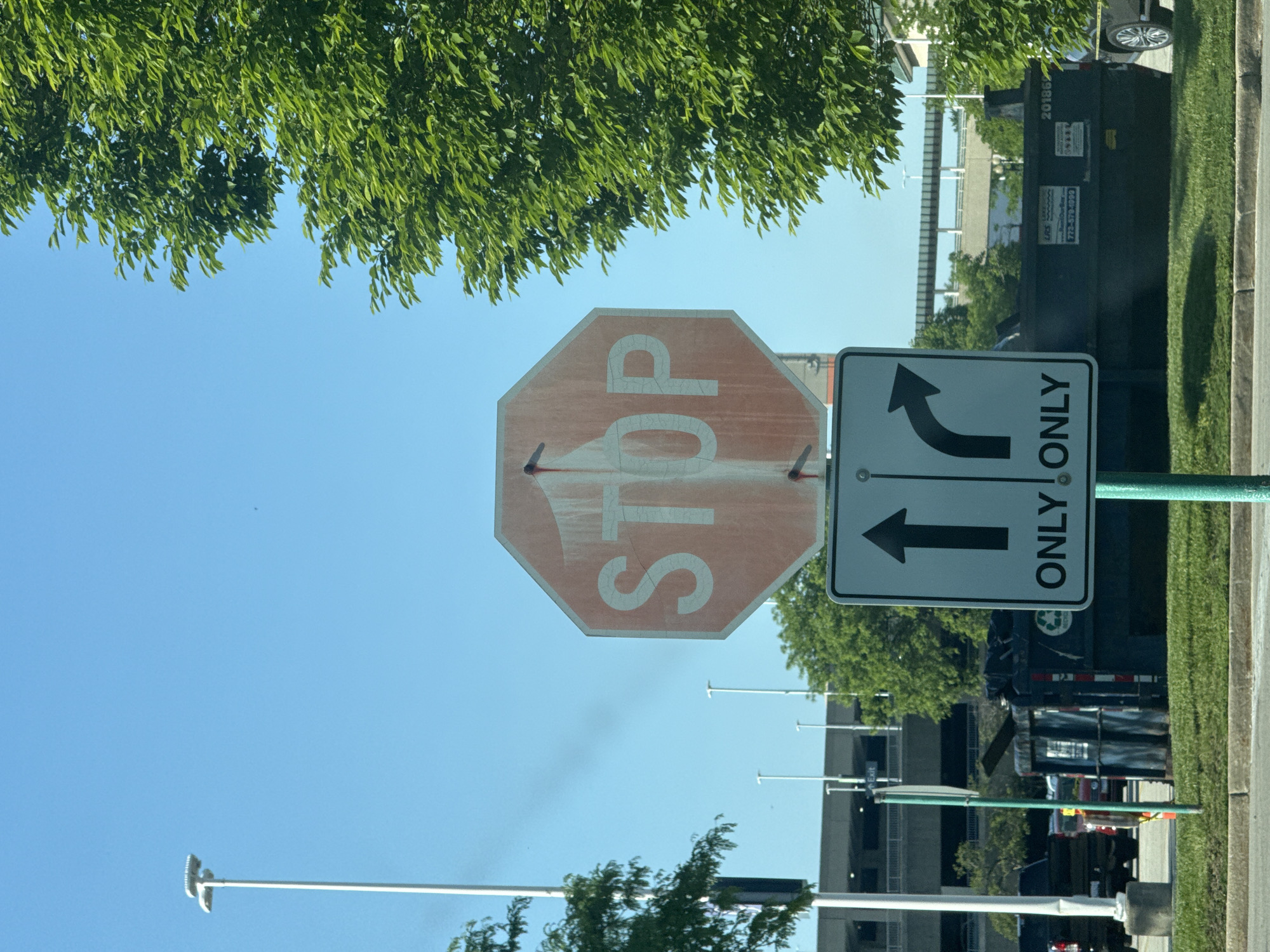 The image depicts a stop sign accompanied by an arrow sign indicating that traffic can only go straight or turn left, located in Skokie, United States of America. The sky is blue, with some green tree foliage visible above and to the sides of the signs. In the background, there is a commercial dumpster, a building, and several light poles. The stop sign appears faded and worn, suggesting it has been exposed to the elements for some time.