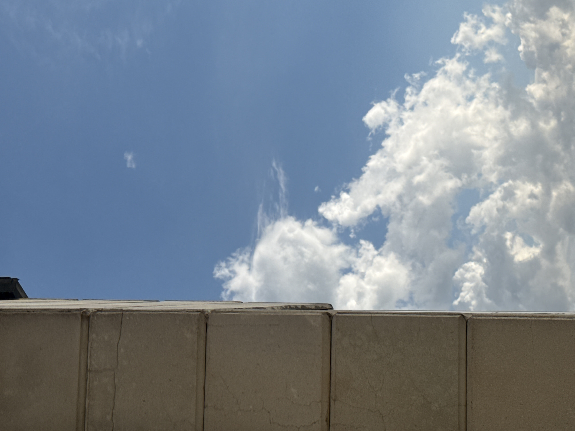 This is a vertical, medium shot photograph taken from below, looking up at the sky from a city street in Abovyan, Armenia. The left side of the frame is dominated by a cream-colored stone building or structure, composed of at least three stacked rectangular blocks. The stone appears aged and shows some signs of cracking. The top edge of the frame reveals a dark, sloped element, possibly part of a roof or overhang.
The right two-thirds of the image is a clear blue sky filled with white, fluffy cumulus clouds. The clouds vary in density, with some appearing wispy and others more substantial. A small, isolated cloud is visible higher up in the sky, to the left of center. The sunlight suggests it is daytime, and the overall impression is one of an outdoor urban environment. No people or animals are visible in the frame.