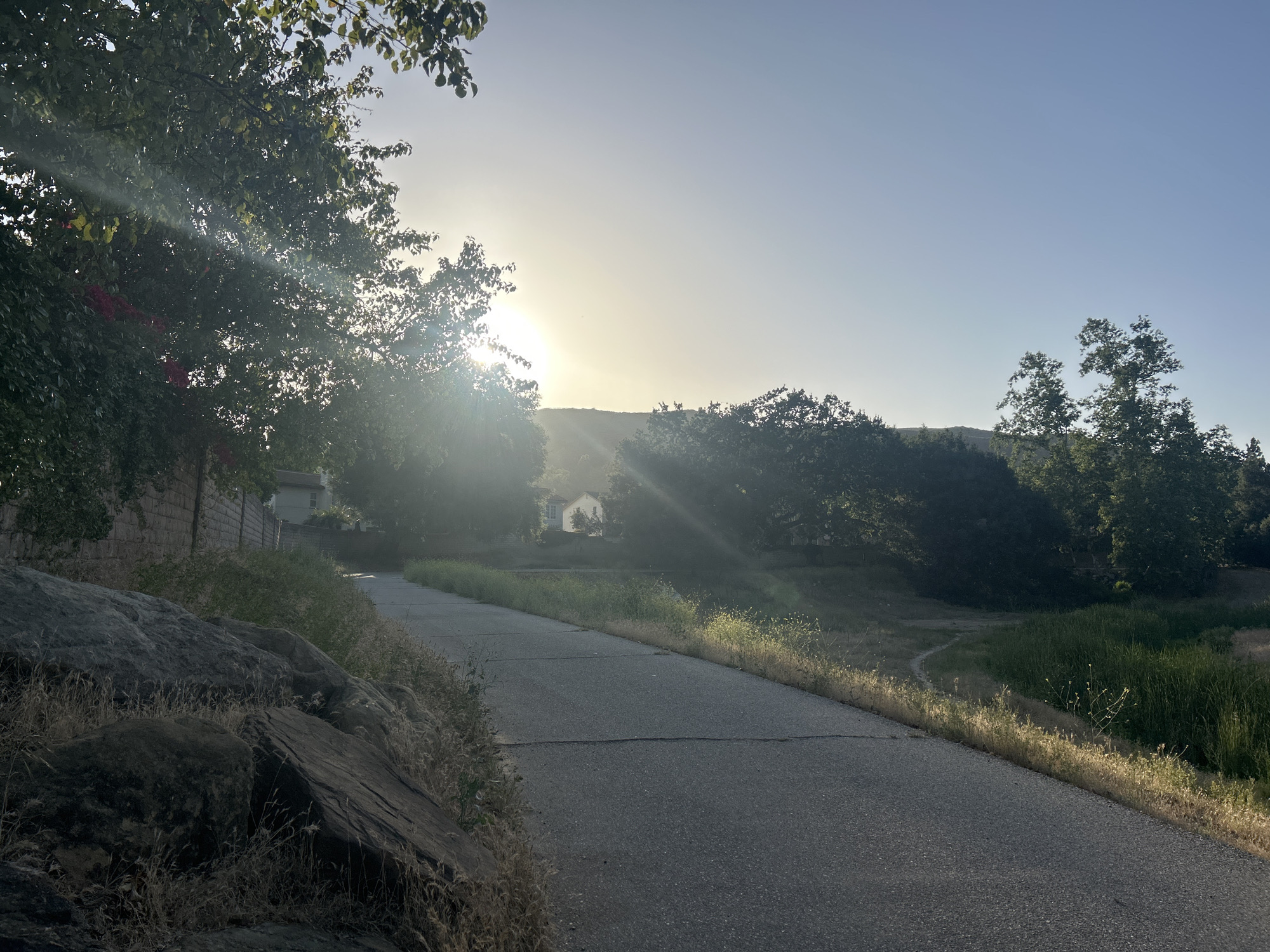 The image depicts a paved road in Oak Park, California, USA, with a large rock formation to the left and vegetation on either side. The sun is visible through the trees, creating a bright, hazy atmosphere. A house is partially visible in the distance, framed by trees. The scene appears to be a walking path or quiet road in a natural or residential area. There are no people visible in the image.