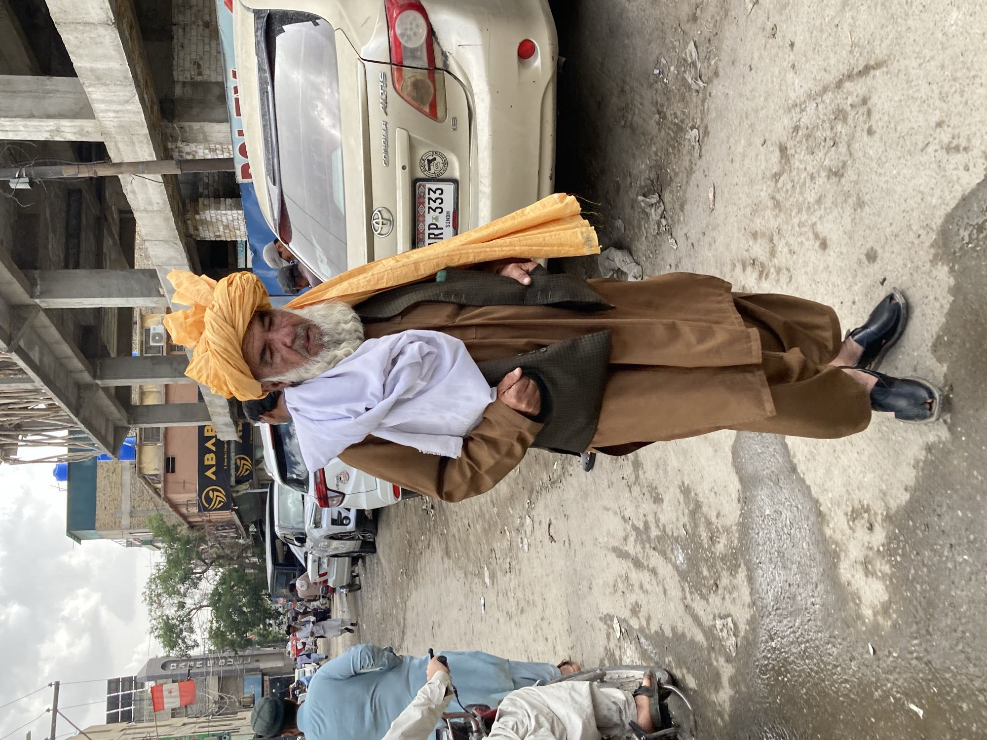 An elderly man stands on the side of a street in Quetta City Tehsil, Pakistan. He wears a brown shalwar kameez, a yellow turban, and a white scarf. Cars are parked nearby, and a crowd of people is visible in the background. The street appears somewhat congested.