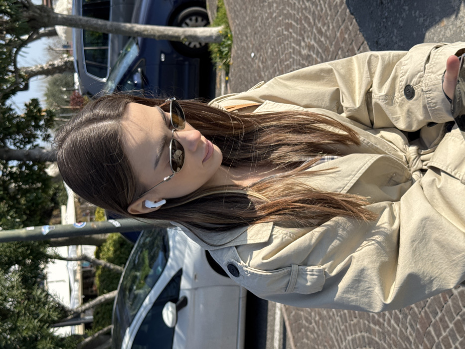 A young female individual is depicted outdoors in Sirmione, Italy, under bright sunlight. Her medium-long brown hair, with lighter highlights, is parted centrally. She wears silver-framed, oval-shaped sunglasses with dark lenses and a white earbud in her right ear. Her attire includes a beige trench coat, belted at the waist, featuring shoulder epaulets and buttoned sleeve straps. A plaid garment is partially visible beneath the trench coat. Her head is angled downwards, with her gaze directed towards a smartphone, partially visible in her hands at the bottom of the frame. The immediate environment includes a paved surface, a dark utility pole, and parked vehicles in the background: a white hatchback on the left and a dark blue sedan on the right. Green foliage from trees is visible behind the vehicles, with faint building structures discernible further in the distance. The scene is illuminated by strong sunlight, casting distinct shadows.