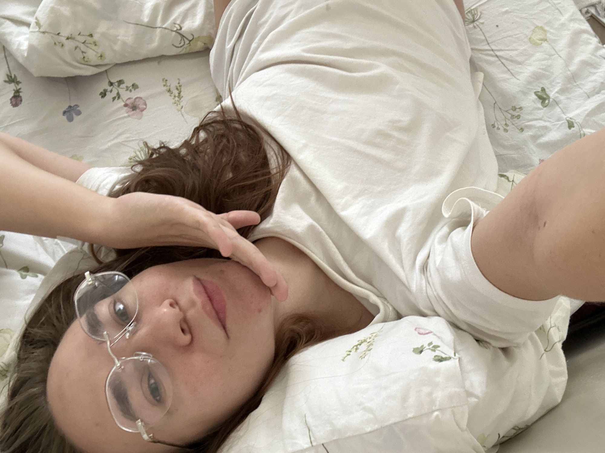 A woman lies on a bed covered with floral print bedding. She wears a white t-shirt and rimless glasses. Her brown hair is spread on the pillow. Her hand is near her face. The location is not discernible as Kharnavy, Russia.