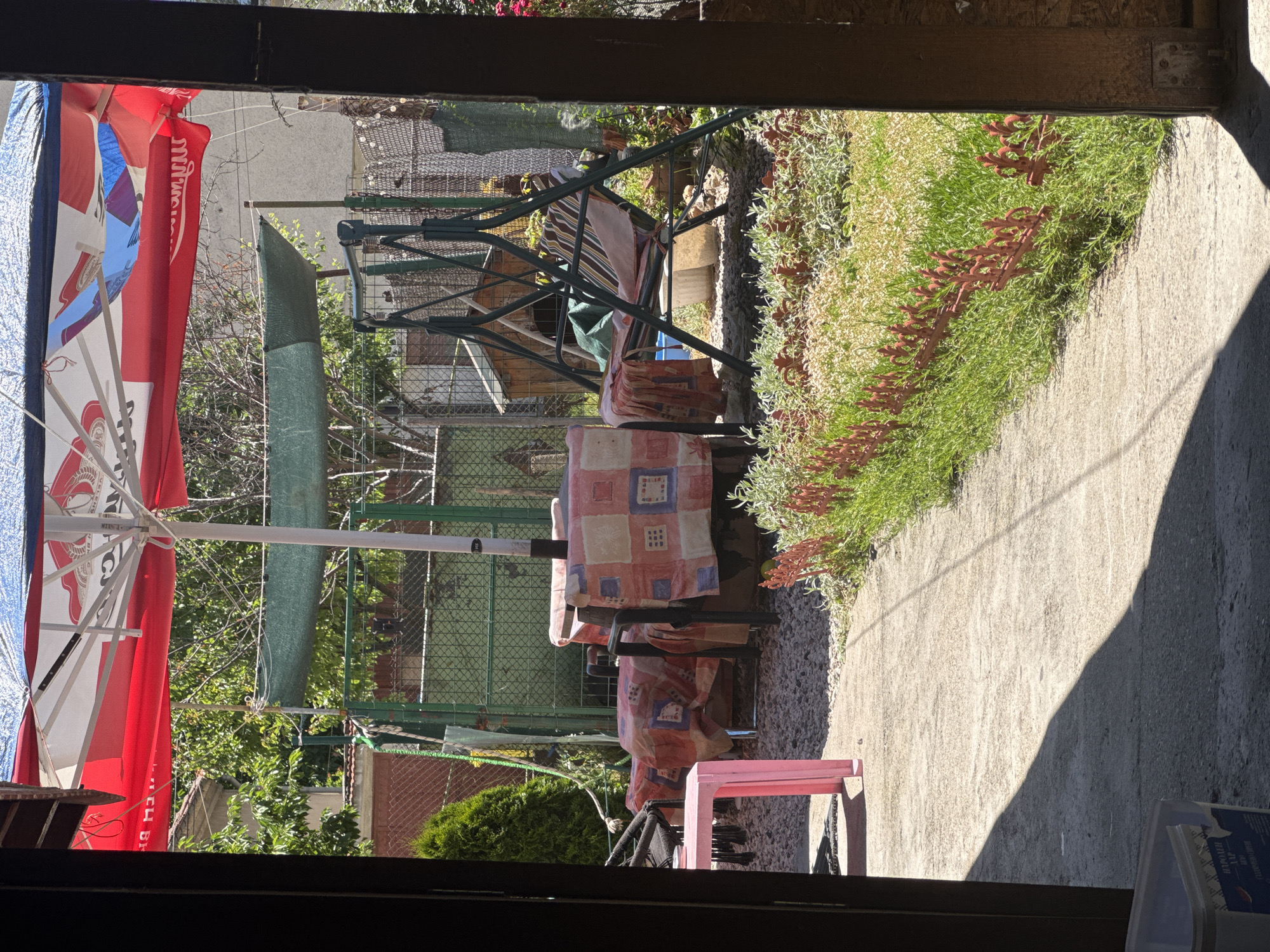 The image depicts an outdoor seating area, likely in a residential backyard, in Svilengrad, Bulgaria. A large red and white umbrella provides shade over a picnic table covered with a patterned cloth. To the right, a green metal swing set is visible, with a striped cushion and several blankets draped over it. A small pink table is located to the left of the picnic table. The ground is covered with gravel and grass, with a concrete path leading into the scene. A wooden dog house is partially visible behind the swing set. A green mesh shade structure is suspended between poles. The background shows fencing and some vegetation. The lighting suggests a sunny day.