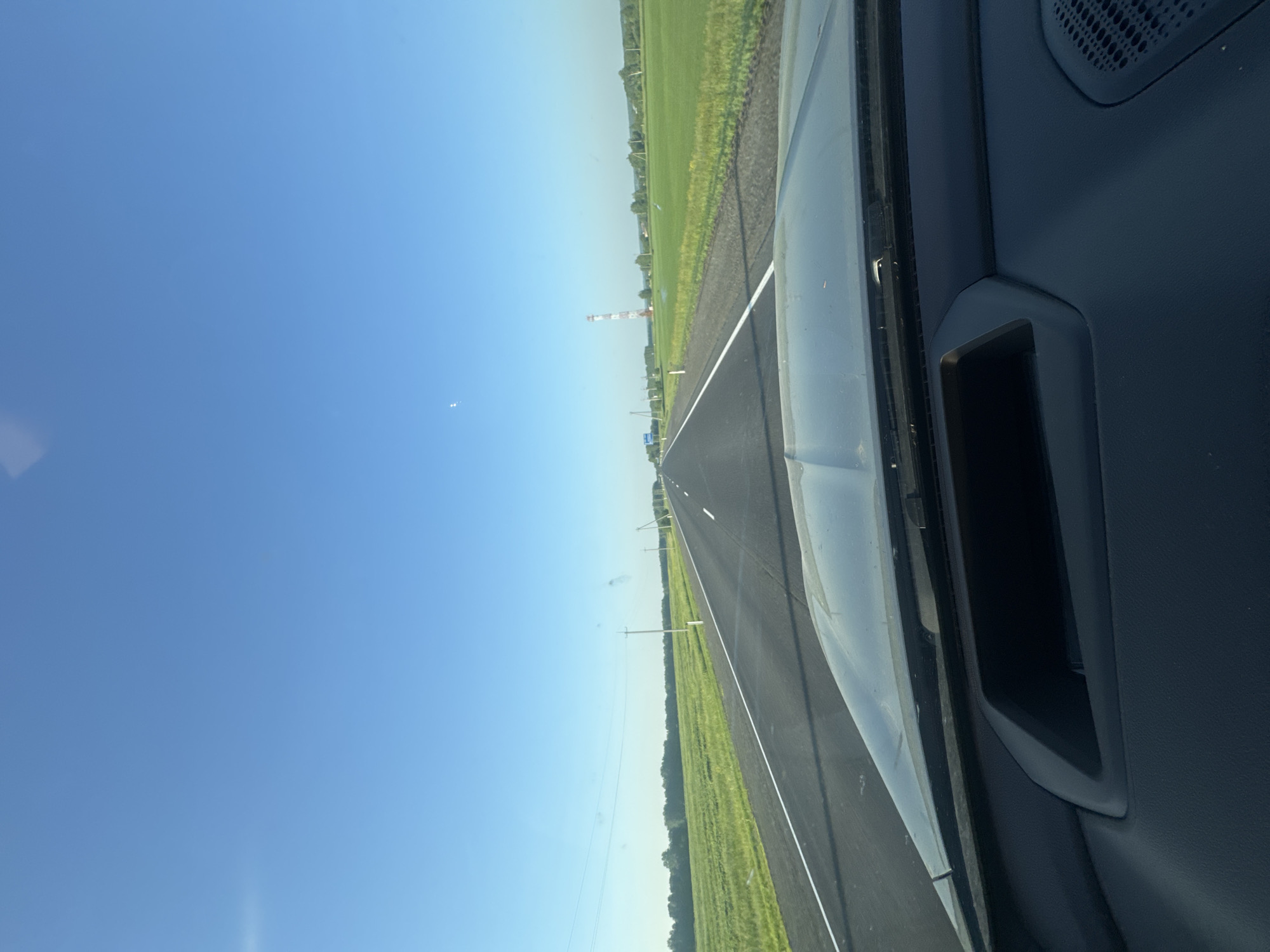 The image shows a view from the inside of a vehicle looking out at a highway on a clear day. The road stretches into the distance, with green fields on either side. There are power lines running along the left side of the road. In the distance, a red and white striped tower is visible. The sky is a clear, bright blue with a few faint clouds. The interior of the vehicle, including the dashboard, is visible in the foreground. The image appears to have been taken in Maltsevo, Russia, given the context provided.