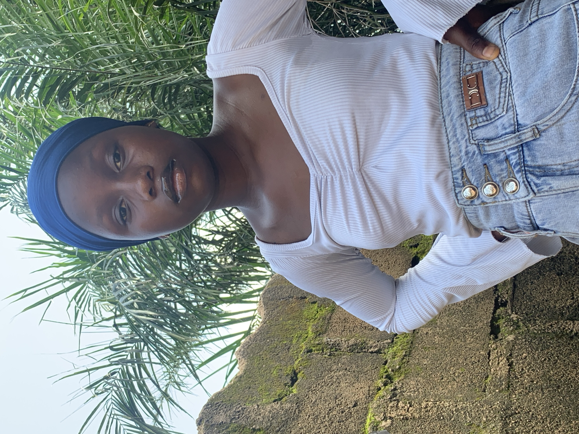 A medium shot shows a young Black woman standing outdoors, looking at the camera. She is wearing a blue headscarf, a white ruched long-sleeved shirt, and light-wash denim jeans. The woman's hands are on her hips, and she appears to be posing. Behind her, there is a moss-covered stone wall and lush green palm fronds. The sky is visible in the upper background. The overall lighting suggests it is daytime. The location is Sabon Tasha, Nigeria.