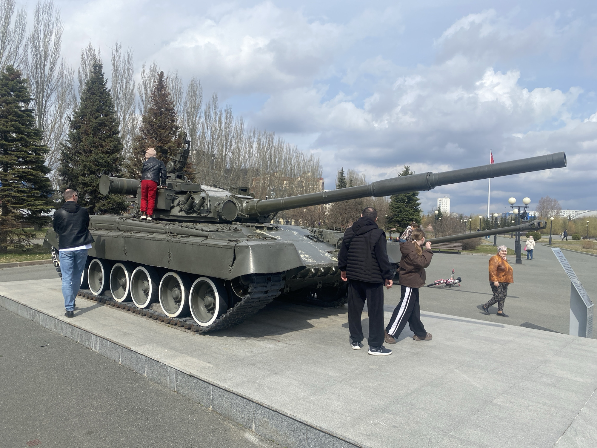 The image displays a large, olive-green main battle tank, likely a T-series, set on a raised grey concrete platform in what appears to be a public park or memorial square in Kazan, Russia. The tank's long barrel extends towards the right, and its track wheels feature distinctive white rings on their rims.
Several people are present in the scene. On the left, a man in a black jacket and blue jeans walks past the tank. A child, wearing red pants, a black jacket, and a light-colored beanie, is climbing or standing on the tank's turret. To the right, two individuals walk away from the tank: one in a black puffer vest and dark pants, and another in a brown jacket and black-and-white striped track pants. Further down the path, an older woman in a brown patterned top and dark leggings walks leisurely. A child's red bicycle lies on its side on the paved path beyond the tank.
The setting features paved walkways, park benches, and streetlights. The background is dominated by a mix of tall, slender deciduous trees, currently bare of leaves, and several dark green evergreen conifers. Distant city buildings are visible through the trees under a bright, partly cloudy sky. A flagpole with a red flag can be seen in the distance. The overall atmosphere suggests a casual daytime visit to a public exhibit or monument. A grey informational plaque stands on the right side of the walkway, but its text is not legible.