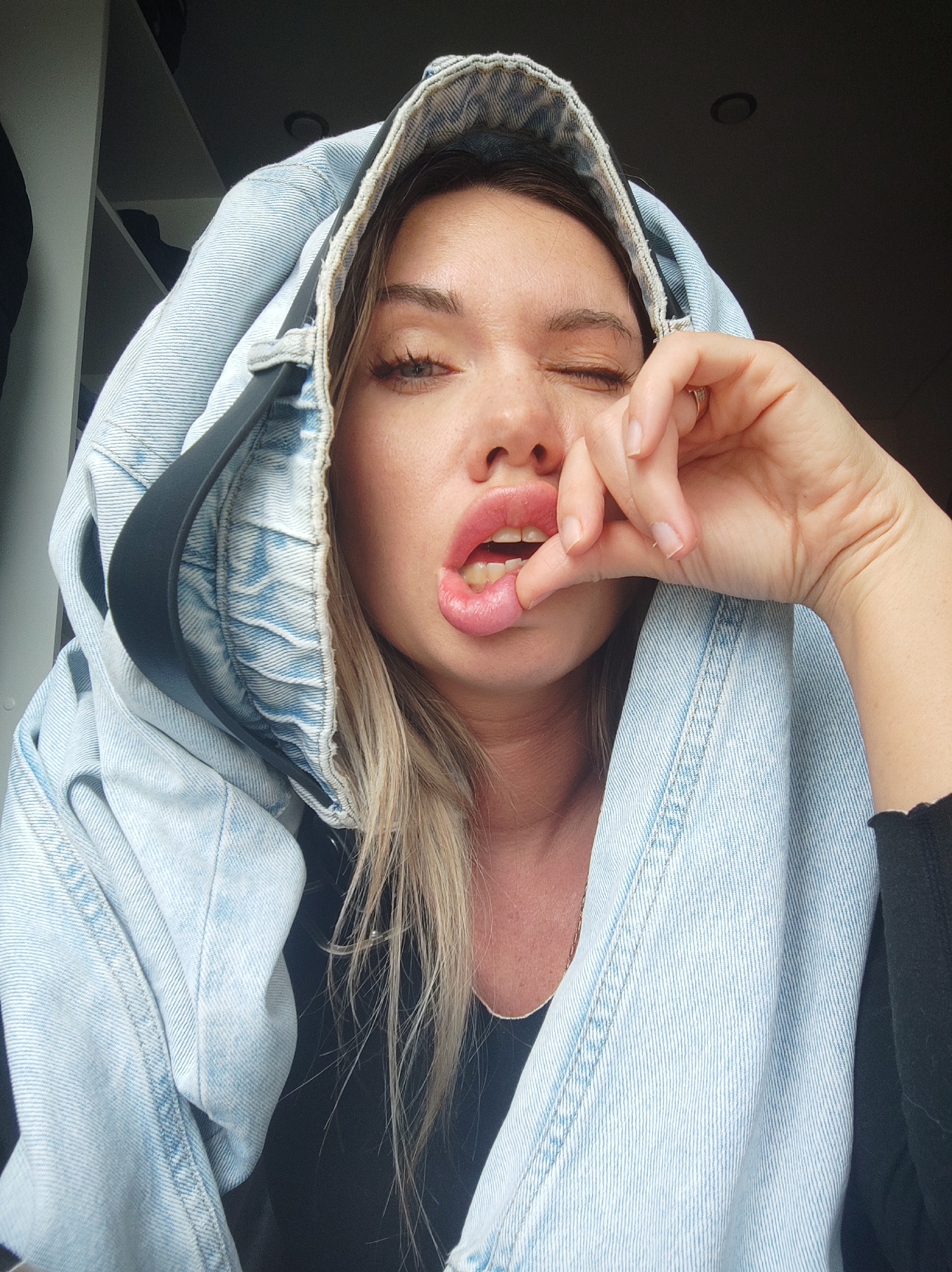A woman with light brown and blonde hair is making a face at the camera. She is wearing a blue denim jacket draped over her head like a hood. Her right eye is squinted closed, and her mouth is open with her fingers pulling at her lower lip. She is wearing a black shirt beneath the jacket. A black strap is visible on the left side of her face, suggesting the jacket might have a shoulder strap. The background is dark, with a shelf partially visible in the upper left corner. There is no indication of the location being Votkinsk, Russia.