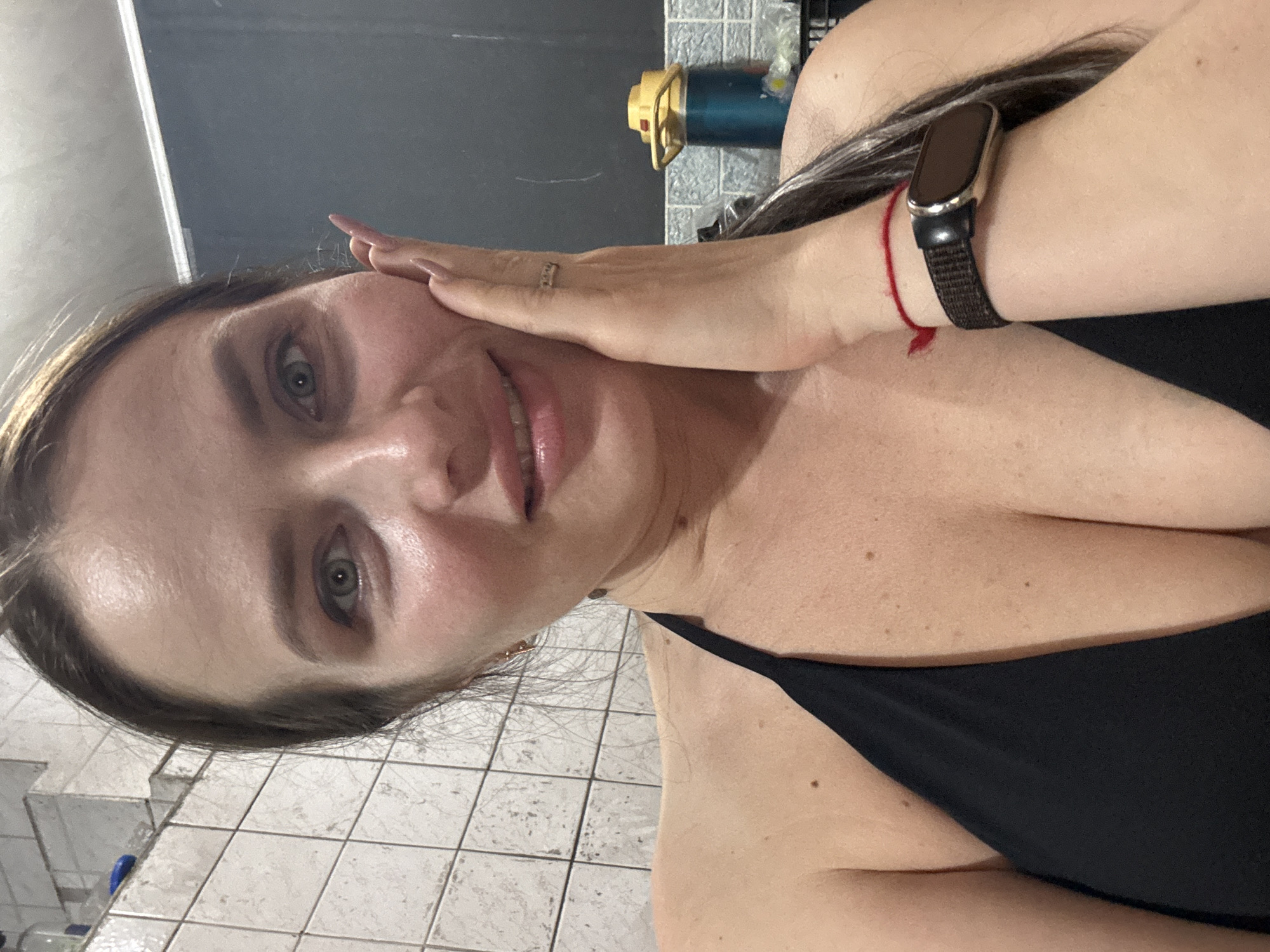 A selfie image shows a woman with light skin, fair hair, and green eyes. She is wearing a black top. Her hand touches her face, and she is wearing a smartwatch on her wrist. The background shows a tiled wall and floor, along with a blurred object on a shelf. There is no information in the image that indicates the location is in Rusalevka, Russia.