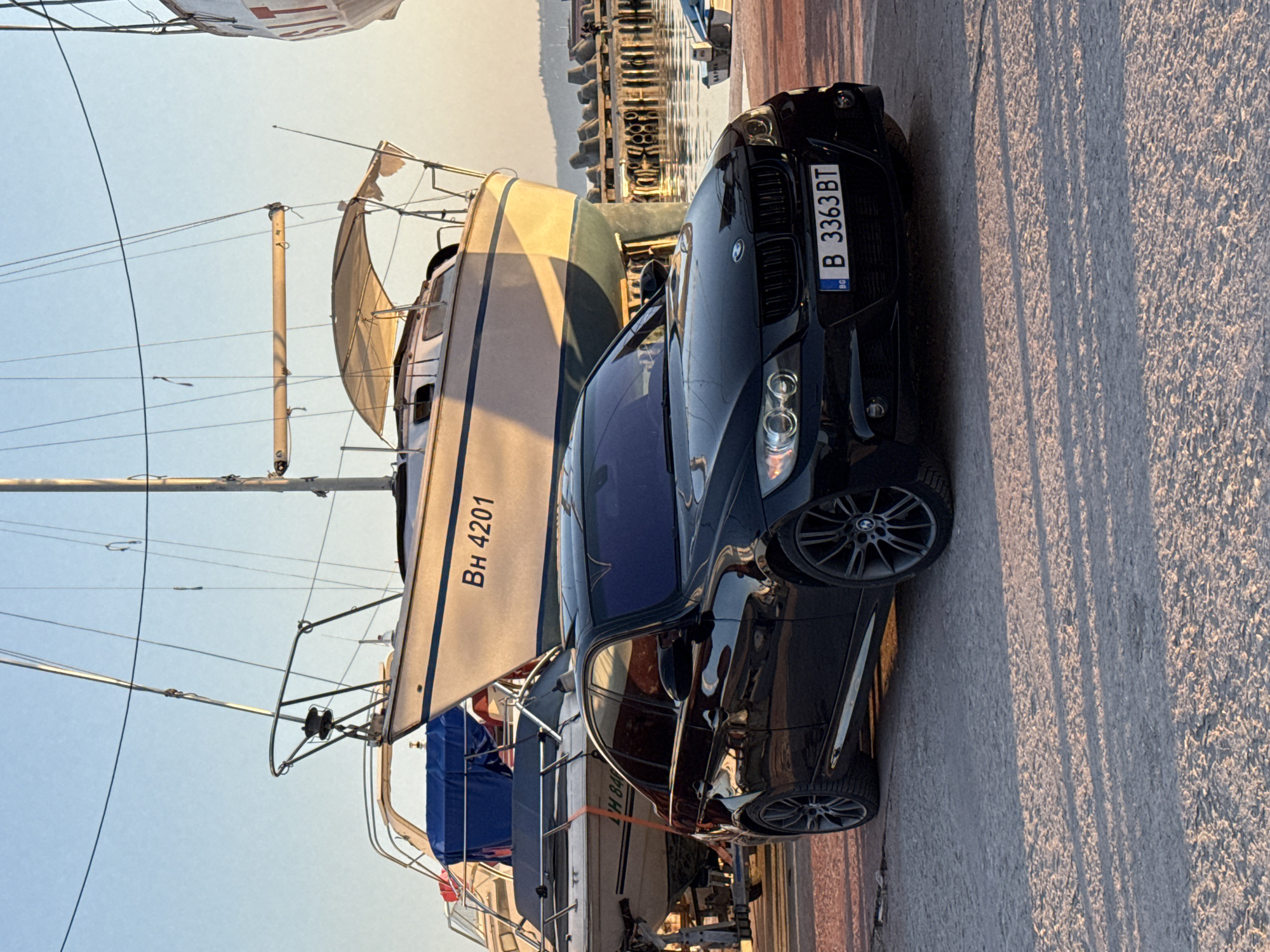 The image shows a black BMW with Bulgarian license plate B 3363 BT parked near the Varna Marina in Varna, Bulgaria. A white sailboat with hull number BH 4201 is docked next to the car. The scene is set during the daytime with clear sky.