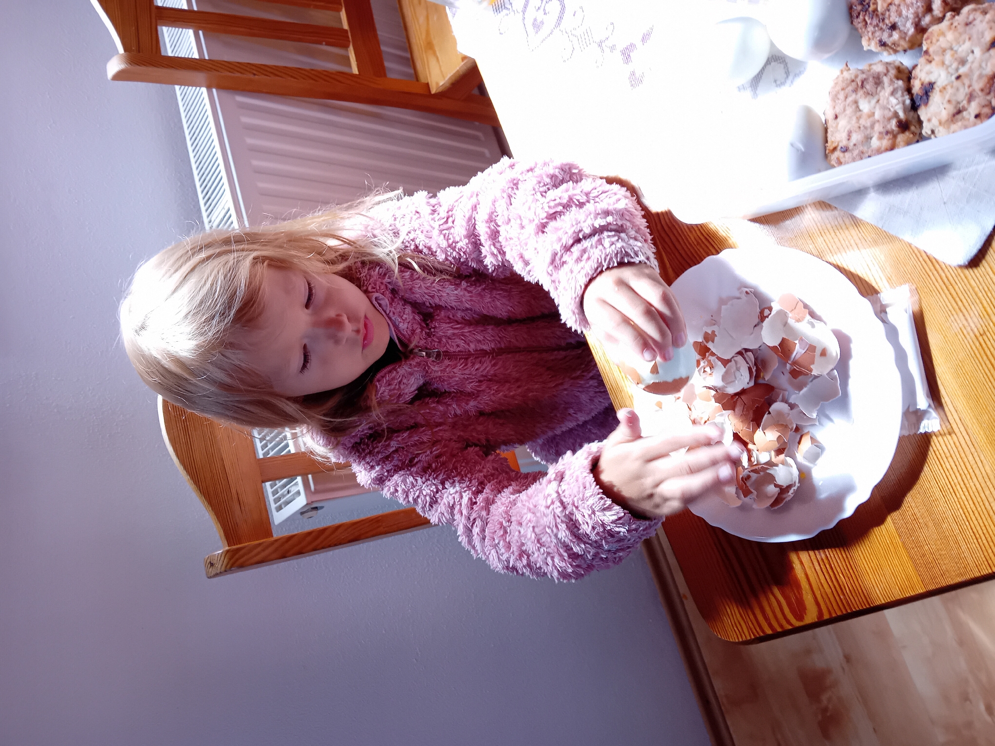 A young girl with blonde hair, wearing a pink fuzzy jacket, is seated at a wooden table and peeling an egg. She is focused on the task, with eggshells piled on a white plate in front of her. To her right, there is a tray containing what appear to be meatballs or small patties, along with two peeled eggs. The background shows a radiator and a wooden chair. The image appears to have been taken indoors, likely in a home setting. The lighting suggests it may be daytime, with natural light coming from the right side of the frame. The location of Zuberec, Slovakia is not discernible from the image itself.
