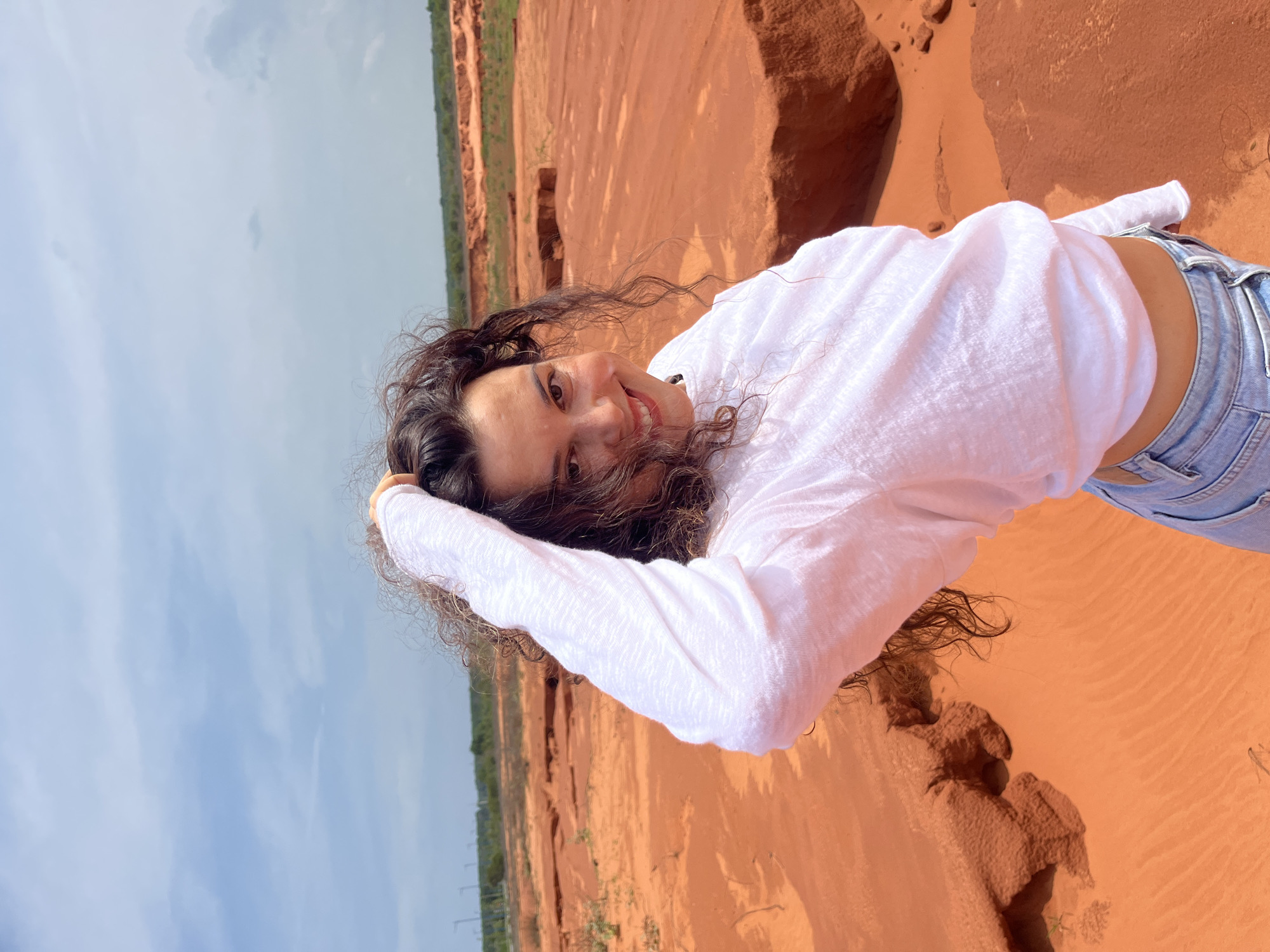 A young woman with curly dark hair and a white long-sleeved shirt is posing in a desolate, sandy landscape with orange-red soil, characteristic of the Red Sand Dunes in Mui Ne, Vietnam. She is smiling at the camera with her left hand raised, touching her hair. The background shows rolling sand formations and a pale blue sky with scattered clouds. In the distance, a line of vegetation and some utility poles are visible. The woman is wearing light blue jeans.