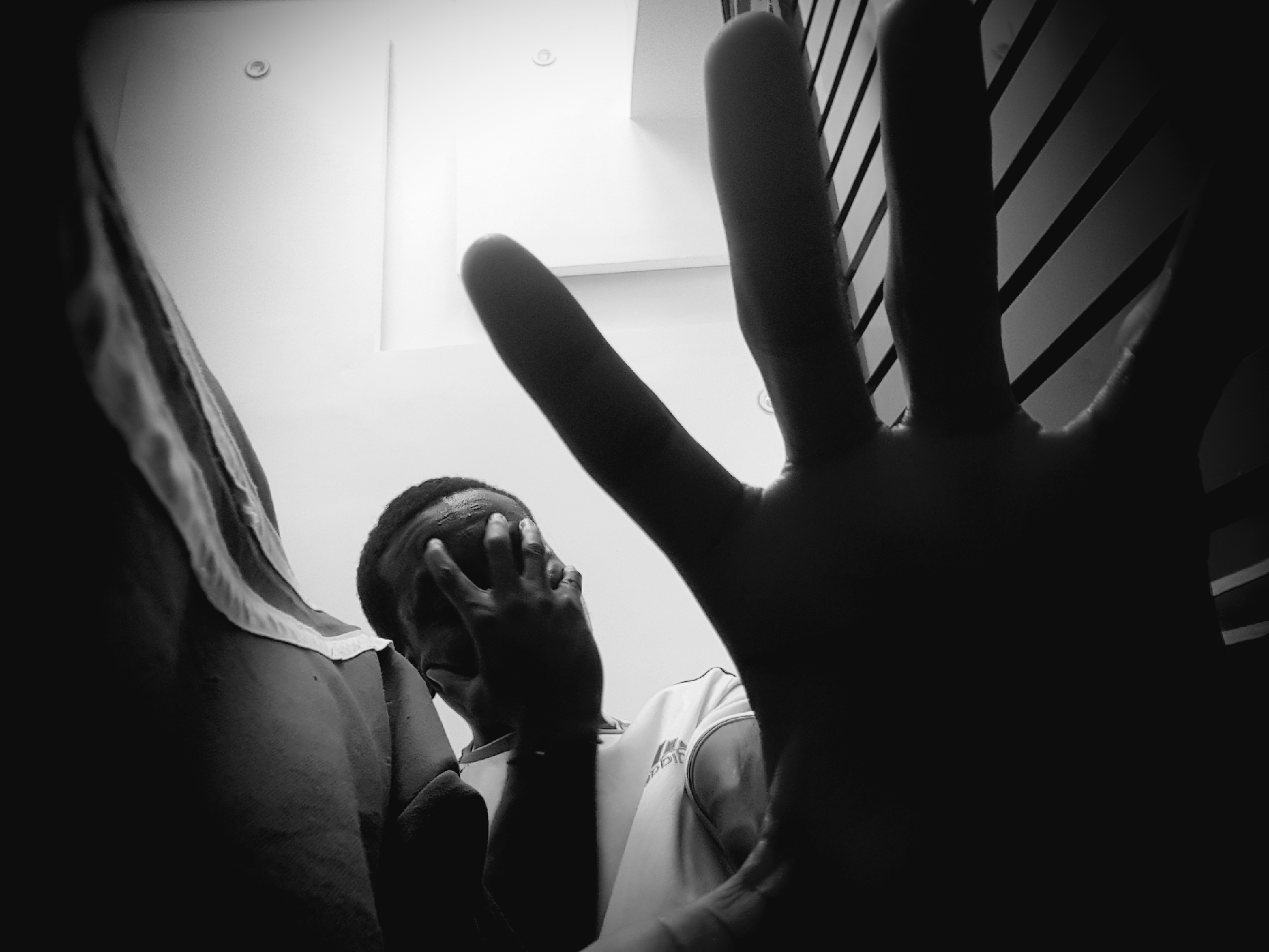 In a low-angle, black and white shot, a person's hands cover their face, head bowed as if in distress or contemplation. The individual, partially visible, wears a t-shirt with an Adidas logo. In the foreground, another hand is outstretched, its fingers blurred by motion or close proximity to the camera, dominating the lower portion of the frame. The background consists of a stark white wall with a circular light fixture and a corner. The scene appears to be indoors, possibly a room, with a dark, ribbed surface, resembling a bed frame or seating, visible beneath the outstretched hand. The lighting creates dramatic shadows, with the left side of the image largely dark and the right side illuminated. The context of Narayi, Nigeria is not visually discernible from the image itself.
