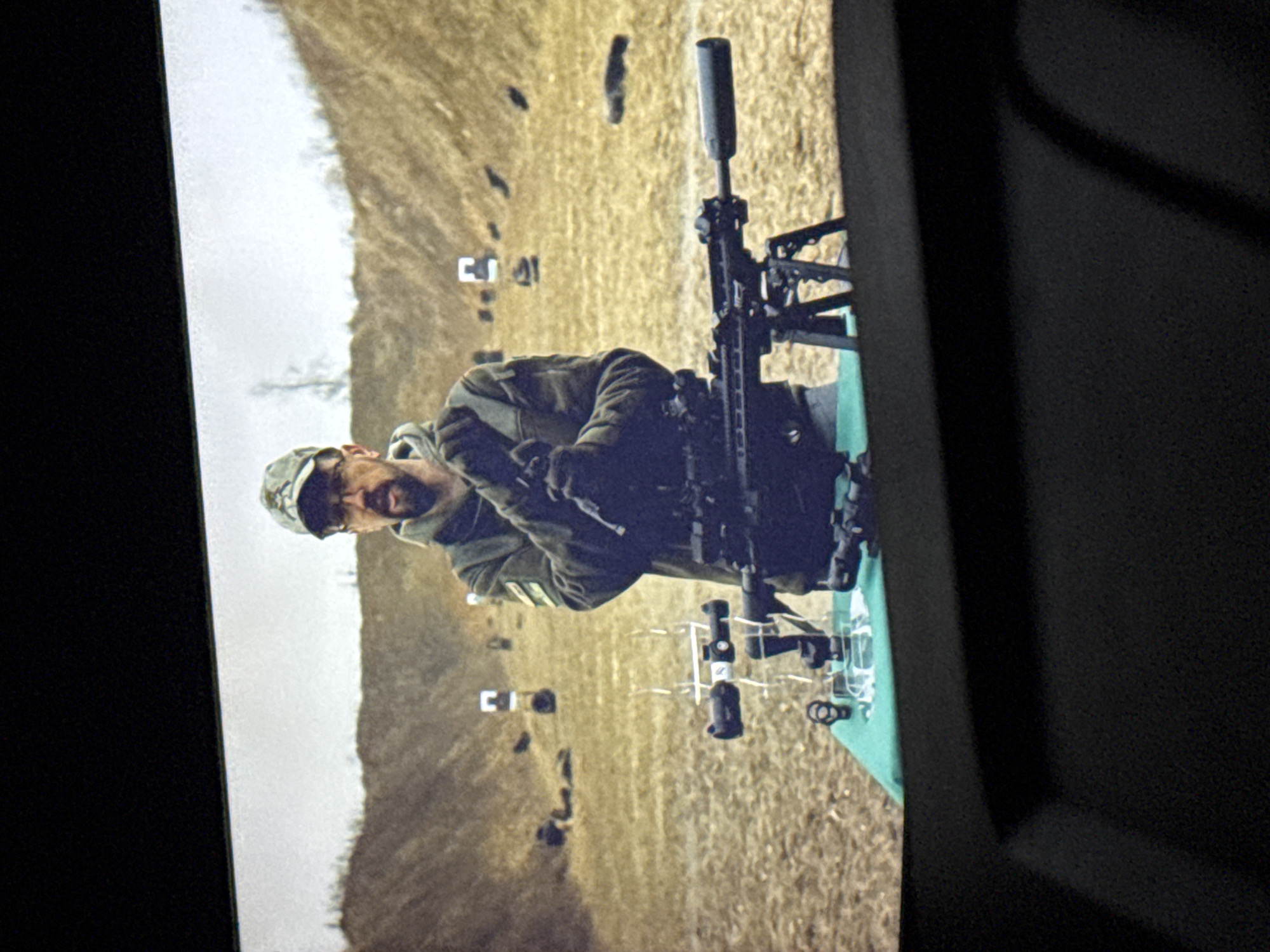 The image shows a man wearing a camouflage cap and glasses, with a beard, standing in front of a rifle range. He is holding a rifle with a suppressor. On a green table in front of him are various optical sights and accessories. The background shows a hill with targets set up at intervals. The scene appears to be a demonstration or training session at an outdoor shooting range. The overall context of Chervonyi Step, Ukraine, suggests this could be related to military training or civilian marksmanship.