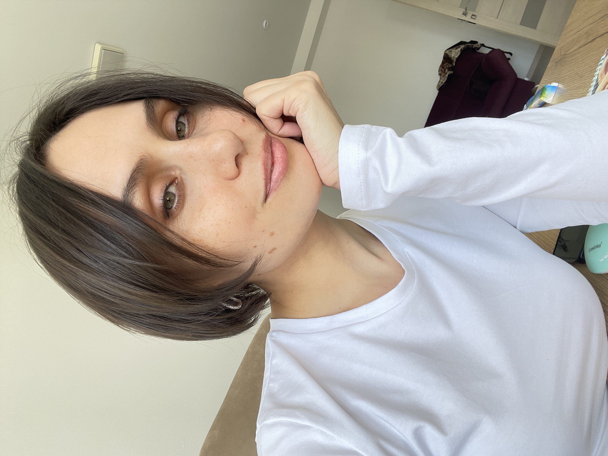 A young woman with short, dark brown hair and light eyes poses for a selfie indoors. She is wearing a plain white long-sleeved t-shirt. Her right hand is touching her face, with her elbow out of frame. In the background, there is a dark burgundy armchair and a glimpse of a wooden surface. The location cannot be determined as Izhevsk, Russia, with the information provided.