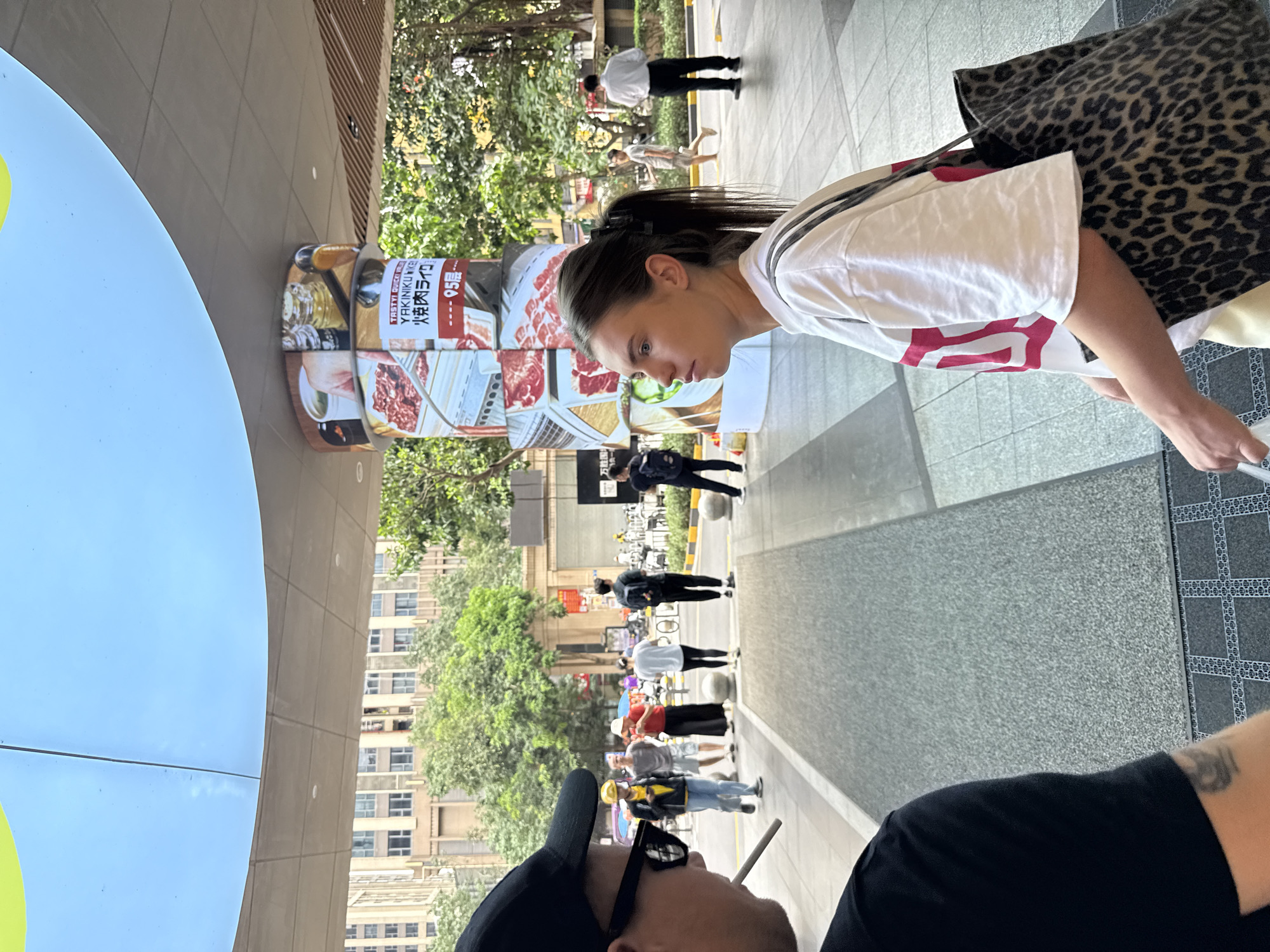In Haizhu District, China, two individuals are in the foreground of the image. The person on the left, seen from the side, wears a black t-shirt, a black baseball cap, and sunglasses, and is smoking a cigarette. The person on the right, a young woman, is facing left, looking towards the side, and is wearing a white t-shirt with red detailing and a leopard-print tote bag. In the background, a cylindrical pillar displays images of raw meat and text, including "TASTY QUICK VALUE YAKINIKU" and Japanese characters. Several other people are walking in the middle ground. The environment appears to be an outdoor urban area, possibly a shopping district, with buildings and trees visible. The ground is paved with textured tiles and concrete.