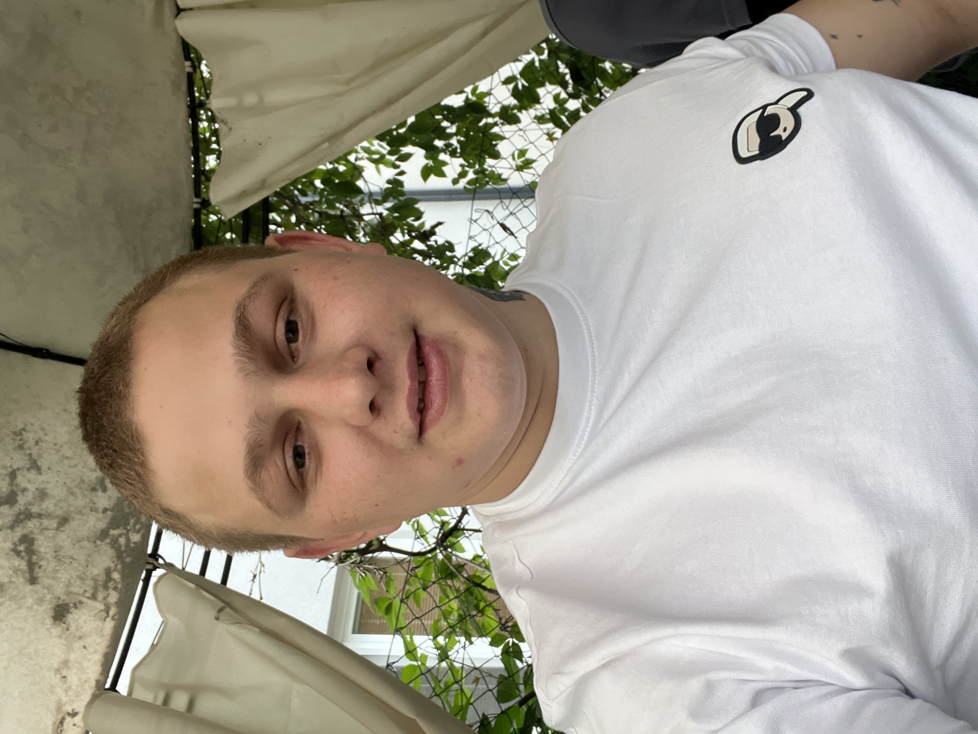A close-up, front-facing selfie shows a fair-skinned individual with a buzz cut, light brown hair, and light brown eyes. The subject is wearing a white t-shirt with a small, stylized cartoon logo on the left chest area. The logo depicts a figure wearing sunglasses and a hat. The subject is outdoors, with green foliage and a chain-link fence visible in the background, suggesting a potentially urban or suburban outdoor setting. The background also includes what appears to be a light-colored canopy or awning overhead. The lighting indicates it is daytime. The image quality is consistent with a smartphone camera. The provided location of Pilsen, Czechia, is not directly evident from the visual information alone.