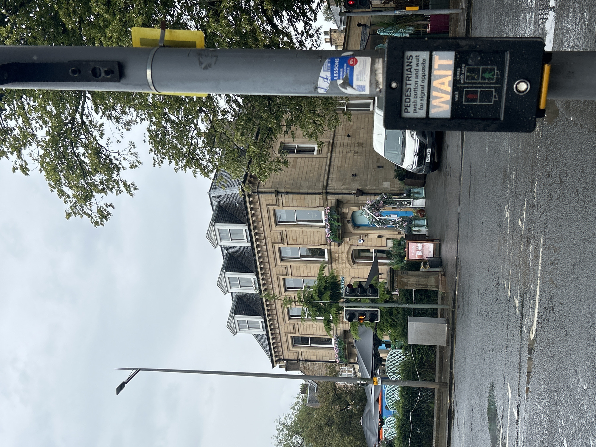The image depicts a street scene in the City of Edinburgh, United Kingdom, on a cloudy day. In the foreground, a pedestrian crossing signal pole stands to the right, displaying the word "WAIT" in illuminated orange letters, indicating that pedestrians should not cross. Below this, illuminated icons show a red standing figure and a green walking figure, with the red figure currently lit. The pole also has a button for pedestrians to press. The street surface is wet, suggesting recent rain, with puddles visible.
To the left of the pole, a traffic light is visible, showing an amber signal. Beyond the street, a building with a stone facade and a dark tiled roof with dormer windows is prominent. The building appears to be a commercial establishment, with outdoor seating areas covered by dark umbrellas visible in front. A white van is parked on the street in front of the building. Trees with green foliage are present, some with branches extending over the building and street. The sky is overcast and grey. A lamppost is also visible to the left of the building.