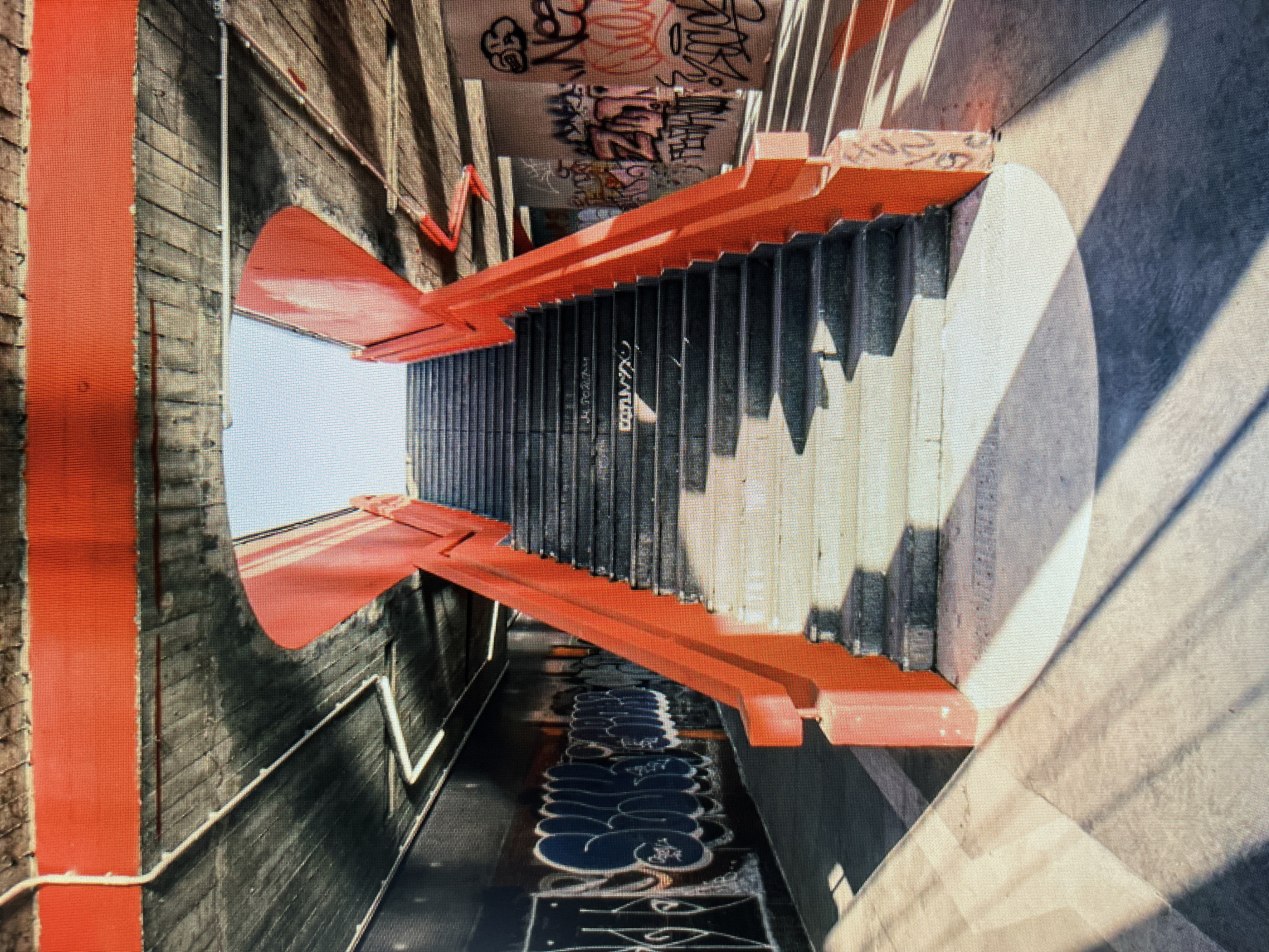 The image depicts an outdoor concrete staircase with bright red railings, leading upwards towards an opening in a ceiling. The staircase is flanked by walls covered in graffiti. Sunlight casts distinct shadows on the steps and the surrounding paved area. The environment appears to be an underpass or a pedestrian walkway. There are no people or animals visible in the image.