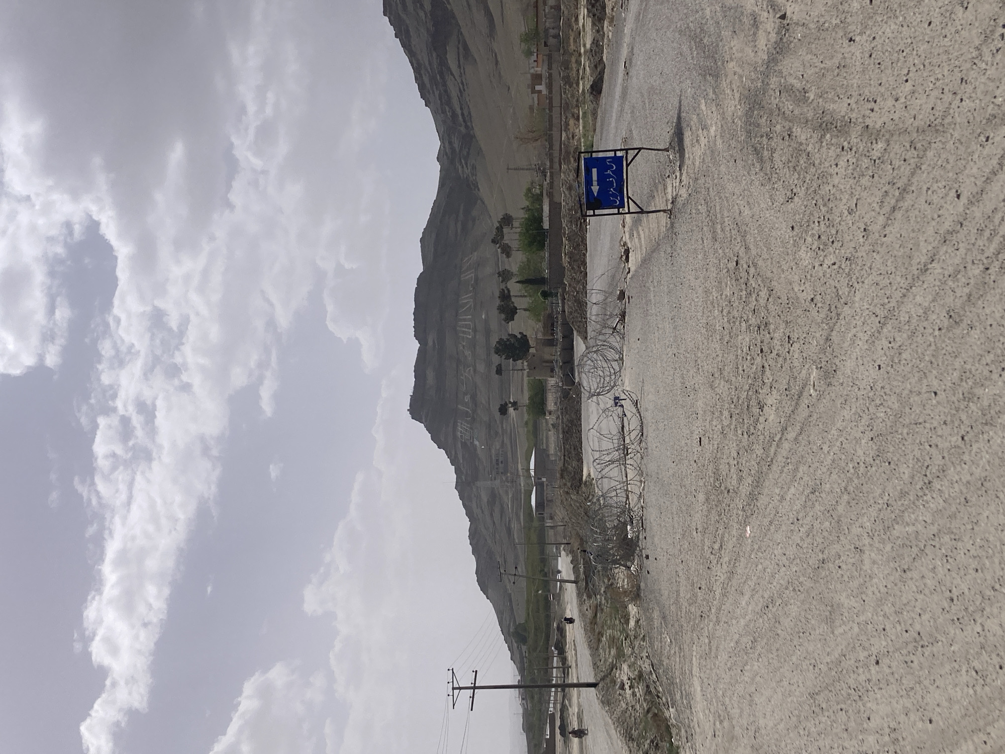 The image depicts a landscape in what appears to be Gulistan, Pakistan, judging by the directional sign written in Urdu. A prominent mountain range rises in the background. The sky is overcast with heavy clouds. A paved road runs through the scene, with a barbed wire fence lining one side. Utility poles and lines are visible along the road. The landscape appears arid. No people are clearly visible in the frame, though there might be some further away. The overall impression is one of a rural or semi-rural environment.