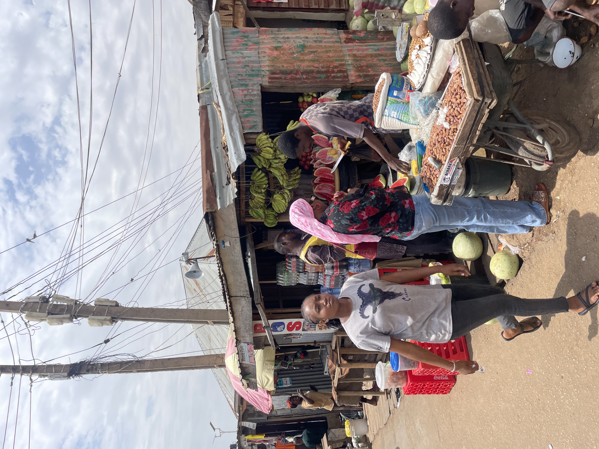 The image depicts a street scene in Jos, Nigeria, with a fruit vendor selling bananas, watermelons, and other produce from a makeshift stall. Several customers are present, including a woman in a grey t-shirt and black pants who is walking towards the camera. The stall is made of corrugated metal and is cluttered with various items, including bags of peanuts. A power pole with tangled wires stands nearby. Coca-Cola crates are visible in the foreground. The street is unpaved.