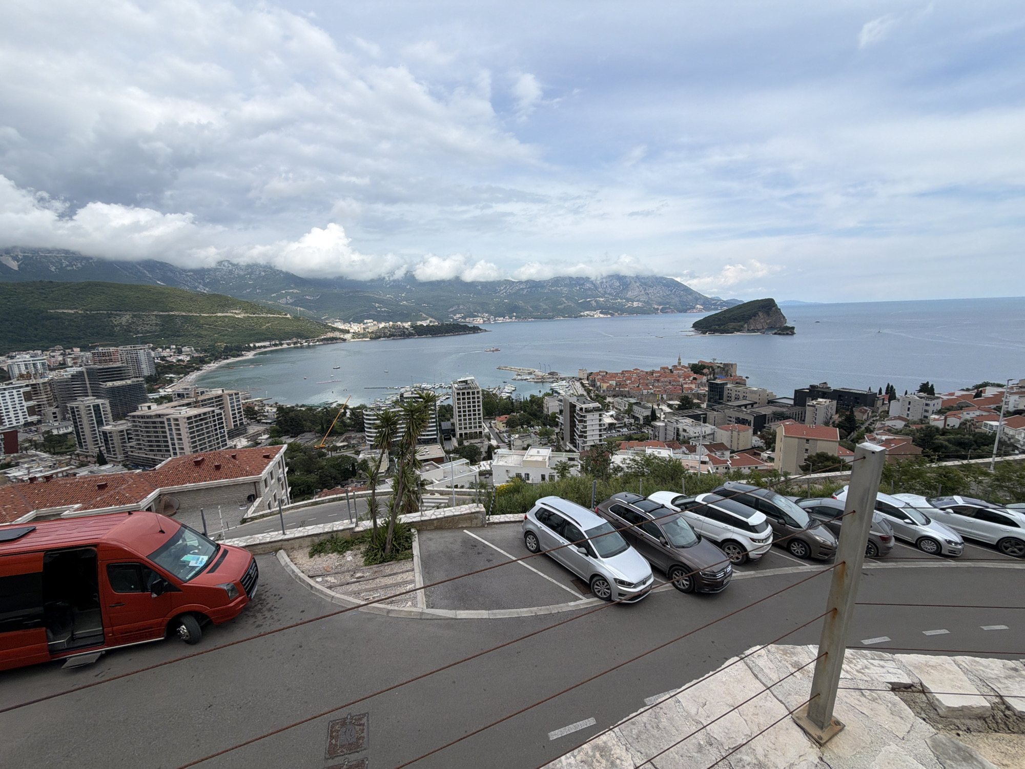 The image captures an elevated perspective of Budva, Montenegro. Visible elements include a red van parked on a road, a row of parked cars, and a cityscape that blends modern high-rise buildings with traditional red-roofed structures. The Adriatic Sea extends in the background, punctuated by an island, possibly Sveti Stefan. Overcast skies with cloud cover complete the scene.