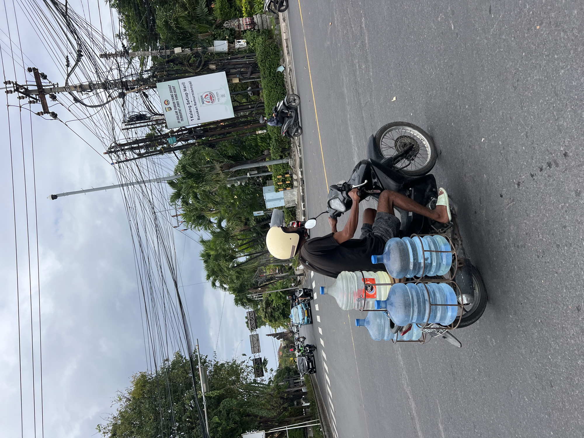 A man wearing a light yellow helmet and a dark t-shirt rides a black motorcycle on a city street. He is carrying five large water jugs on the back of the motorcycle, three are blue, and two are clear. The motorcycle is in motion. In the background, there are other vehicles on the road, palm trees, and a sign with Indonesian writing. The sky is overcast. The location is the intersection of Jalan Hayam Wuruk and Jalan Gajah Mada in Denpasar, Indonesia.